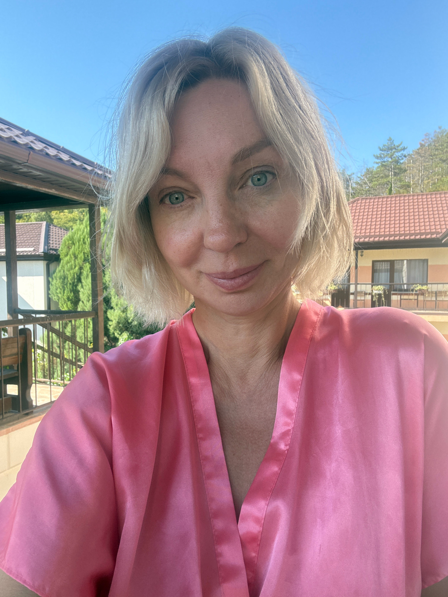 A blonde woman with blue-green eyes smiles at the camera, wearing a pink satin robe. She is standing on a balcony or terrace with a railing. In the background, there is a building with a red-tiled roof, trees, and a clear blue sky. The location appears to be the city of Abrau-Dyurso, Russia.