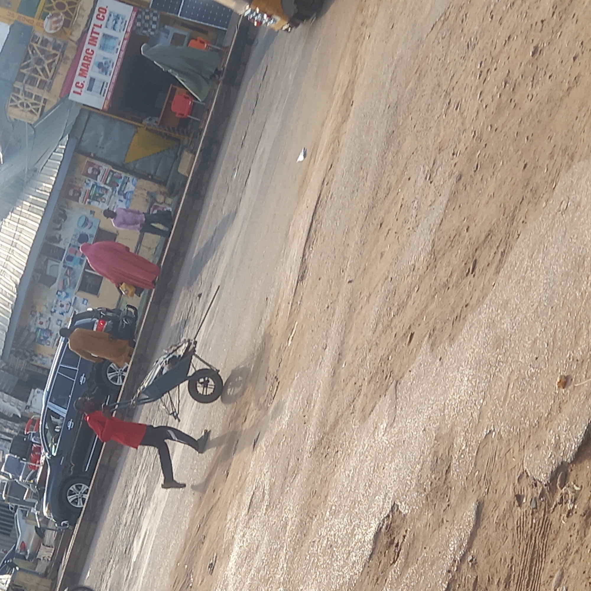 The image depicts a street scene in Dadi Riba, Nigeria. A man in a red shirt and dark pants is pulling a wheelbarrow along the side of a street. Several people dressed in traditional Nigerian clothing are visible near a dark car parked by the side of the road. A building with a sign that reads "I.C. MARC INTL. CO." is situated along the edge of the street. The street surface is rough, appearing to be a mix of dirt and pavement.