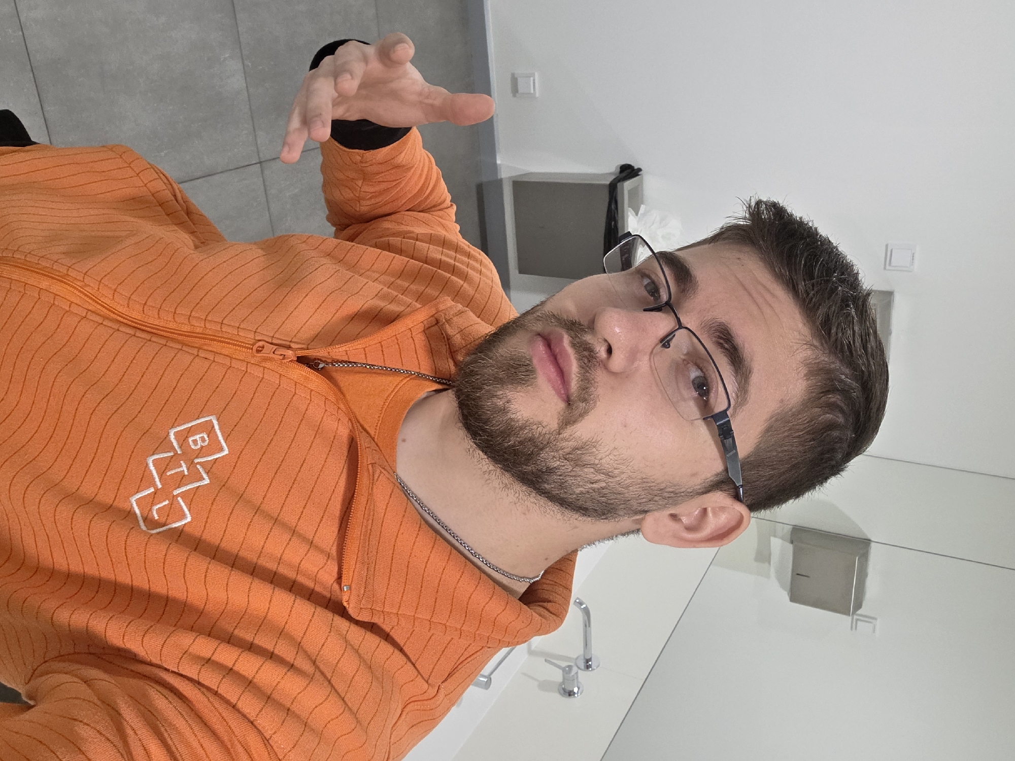 A light-skinned adult male with short brown hair, a trimmed beard, and black-framed eyeglasses is depicted in a medium shot, looking directly at the camera. He is wearing an orange long-sleeved zip-up top with subtle horizontal ribbing and a white "BTL" logo, consisting of three outlined squares, on the left chest. An orange t-shirt is visible beneath the zip-up, and a silver chain necklace is around his neck. His left hand is extended towards the viewer, palm open and fingers slightly bent, positioned approximately at his chest level.
The scene takes place indoors, likely within a public restroom or similar utility area, characterized by clean white walls and a grey tiled floor. Visible in the background are a stainless steel paper towel dispenser mounted on the upper right wall, a grey waste bin with a black liner on the left, and a chrome faucet and mixer handle on the right, suggesting a sink is present out of frame. White electrical outlets or light switches are also visible on the walls. The precise location within the city of Plovdiv, Bulgaria, cannot be ascertained from the visual evidence.