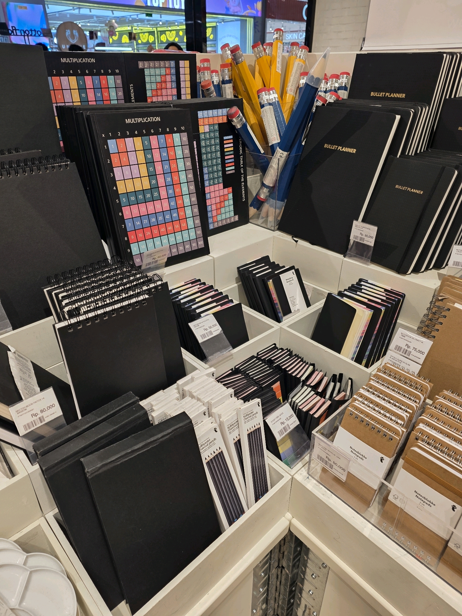 The image depicts a retail store display featuring a variety of stationery items, primarily notebooks and pencils. The setting appears to be an aisle within a larger store, with multiple shelves and display boxes filled with merchandise. Prominent items include black notebooks labeled "BULLET PLANNER," colorful multiplication tables and periodic tables, and a collection of oversized pencils in yellow and blue. There are also smaller notebooks with various colored page edges and spiral-bound sketchbooks. Prices are visible on some of the items, indicated in Rupiah (Rp.). The background suggests a brightly lit commercial environment, possibly a department store or a dedicated stationery shop. No people are clearly visible in the foreground or mid-ground, though a blurry figure might be present in the distant background. The overall impression is one of organized retail display of study and organizational tools. Given the Indonesian Rupiah currency indicated on the price tags, and the typical branding of such stores, this scene is likely from a well-known bookstore chain or stationery store located within a shopping mall in Bandung City, Indonesia.