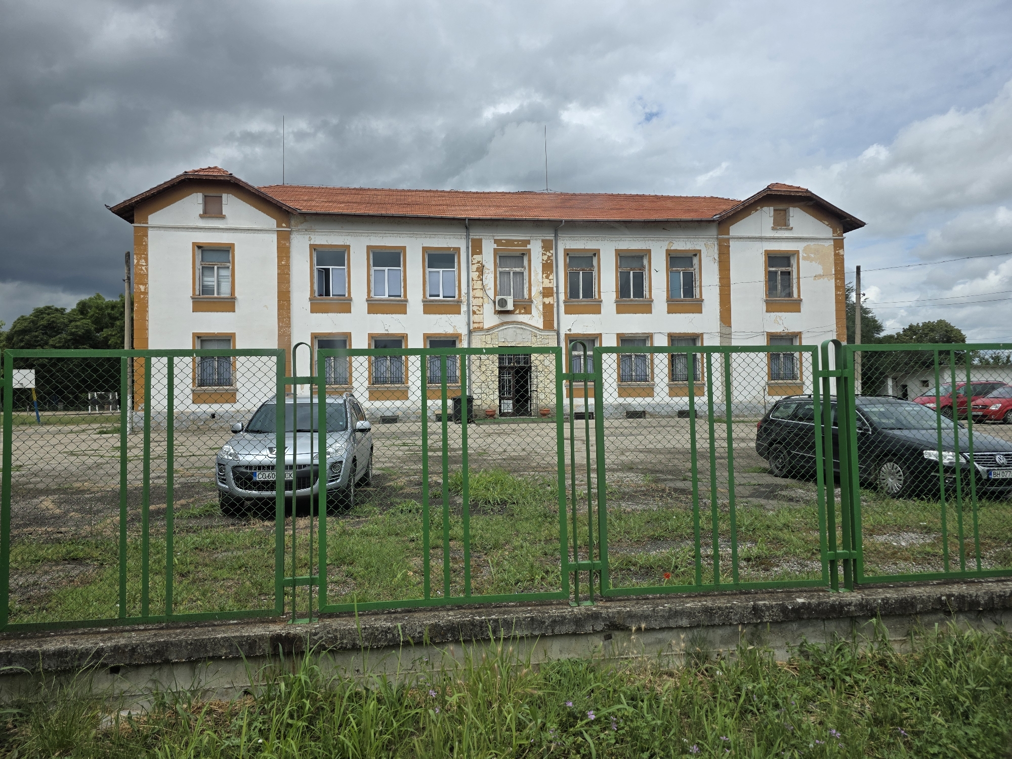 An exterior, eye-level shot captures a two-story building in Dunavtsi, Bulgaria, on an overcast day. The building features a white facade with ochre-yellow trim around its numerous rectangular windows and corners, and under its red-tiled roof. The paint appears aged and peeling in many areas, indicating weathering or lack of recent maintenance. Two prominent gable ends flank the symmetrical structure. A small, external air conditioning unit is mounted above the central entrance.
In the foreground, a green metal fence, comprising both chain-link mesh and vertical bars, runs across the entire width of the image, separating the viewer from the building. The ground inside the fenced area is covered in unkempt grass and some gravel or dirt patches. Two cars are parked within this enclosure: a silver SUV (possibly a Peugeot) on the left, displaying a license plate that reads "CQ 60 CK", and a black wagon (likely a Volkswagen Passat) on the right, with a partially visible license plate starting "BH 027".
The sky is heavily clouded with dark grey formations, particularly towards the left, hinting at impending rain or stormy weather. Trees are visible behind the building on the left side, and utility poles and wires are faintly visible on the right against a slightly brighter, but still cloudy, sky. The overall scene depicts a somewhat neglected yet structurally sound building under a dramatic sky. No specific activity or people are visible.