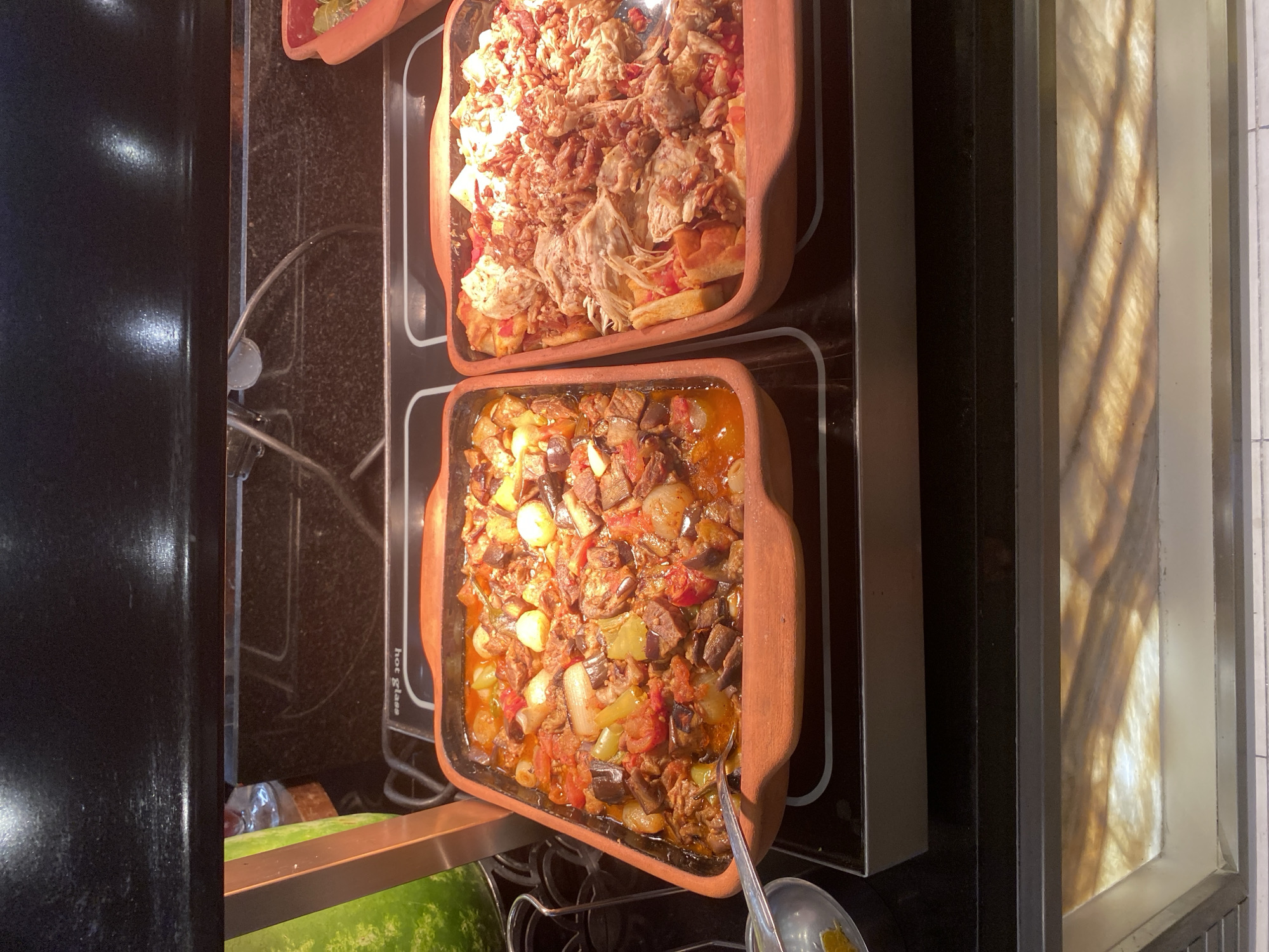The image depicts a buffet-style food service, likely in a restaurant or hotel in Aksu, Turkey. Two clay dishes filled with food are prominently displayed on a heated surface. The dish on the left contains a stew-like mixture of vegetables, including eggplant, tomatoes, and onions, in a reddish-brown sauce. A silver serving spoon rests in this dish. The dish on the right appears to contain shredded meat, possibly chicken or lamb, mixed with bread and other ingredients, also in a saucy base. The surrounding environment shows a dark countertop with a reflective surface and a blurred green object to the left, which might be a beverage dispenser. The lighting suggests an indoor setting, possibly during daylight hours given the warm illumination. The overall scene is focused on the presentation and availability of food.