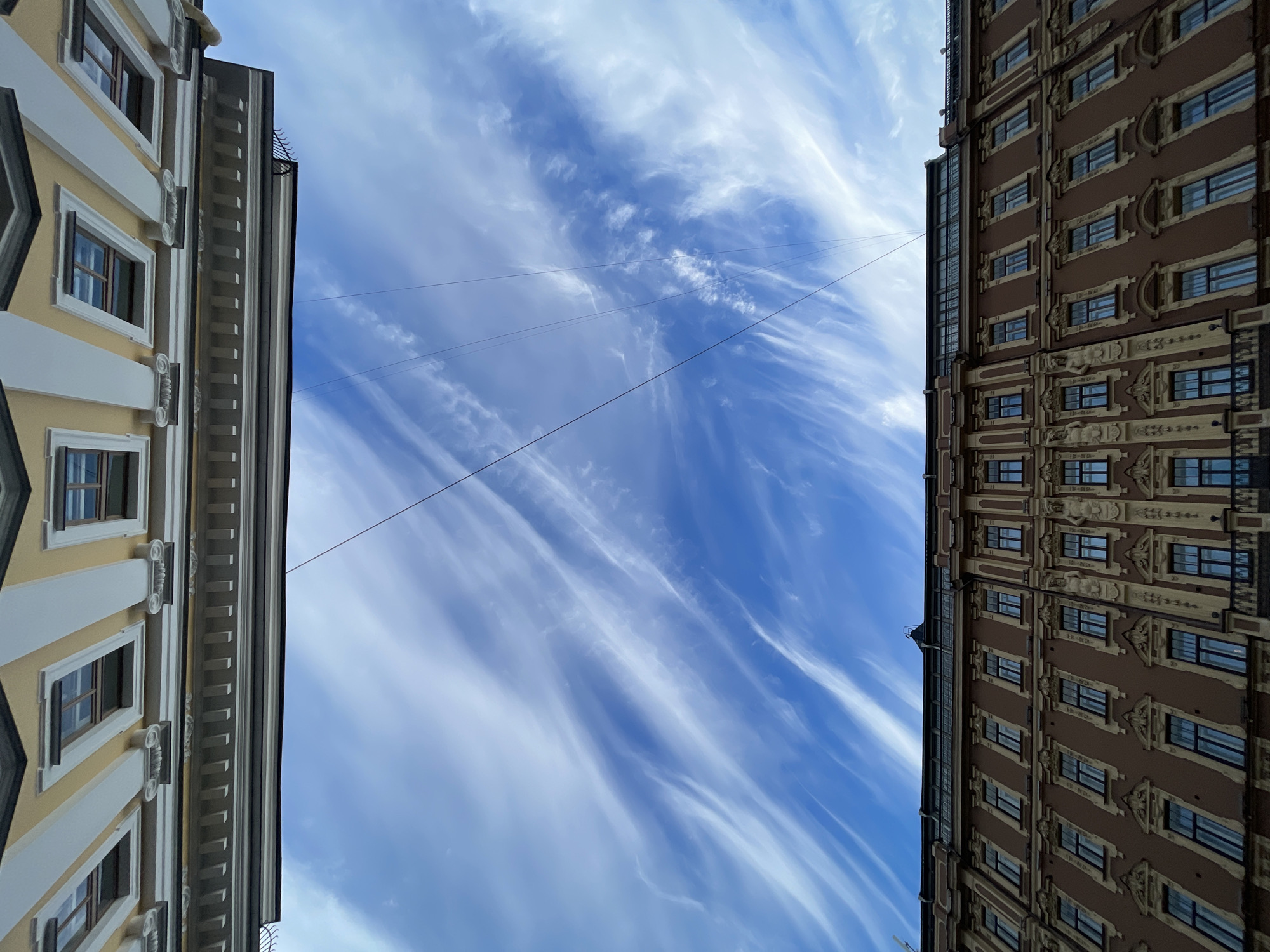 The image presents a low-angle, upward view of a vibrant blue sky framed by two ornate multi-story buildings, suggesting a narrow street or urban passage.
On the left, a building with a light yellow or cream-colored facade rises upwards. It features numerous rectangular windows with dark frames, each set within white decorative surrounds. White pilasters and scroll-like architectural details adorn the spaces between the windows on different levels, giving the building a neoclassical or classical revival appearance. A distinct stepped cornice is visible along its top edge.
On the right, a multi-story building with a reddish-brown facade stands tall. This building is richly ornamented with lighter-colored decorative elements, likely in beige or light stone. These include intricate carvings, figures (possibly caryatids or atlantes) integrated into the architecture on the lower visible floors, and ornate window surrounds. The overall style is an elaborate historicist design, potentially incorporating Art Nouveau elements.
The central part of the frame is filled with a bright blue sky, streaked with long, wispy white cirrus clouds that stretch diagonally across the frame, indicative of high-altitude winds. Several thin, dark utility wires crisscross the sky between the two buildings.
The scene depicts a clear, sunny daytime, suggesting pleasant weather. The distinct and elaborate architectural styles of both buildings are characteristic of historic city centers, consistent with the provided location context of Saint Petersburg, Russia. No people or visible text are present in the image.
