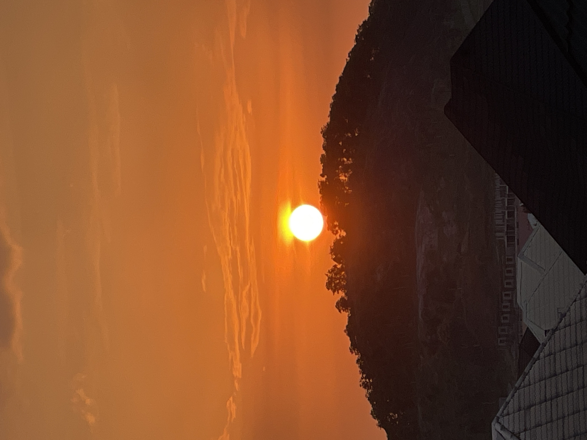 A vibrant orange sunset casts a warm glow over the landscape. The sun, a bright orb, sits just above a silhouetted hilltop covered in trees. In the foreground, rooftops of houses are visible, some tiled and others dark and pointed. The scene appears to be set in a residential area on the outskirts of Abuja, Nigeria, possibly during the golden hour. The sky is filled with scattered clouds, tinged with orange and yellow hues from the setting sun. The overall atmosphere is serene and picturesque, capturing a moment of natural beauty.