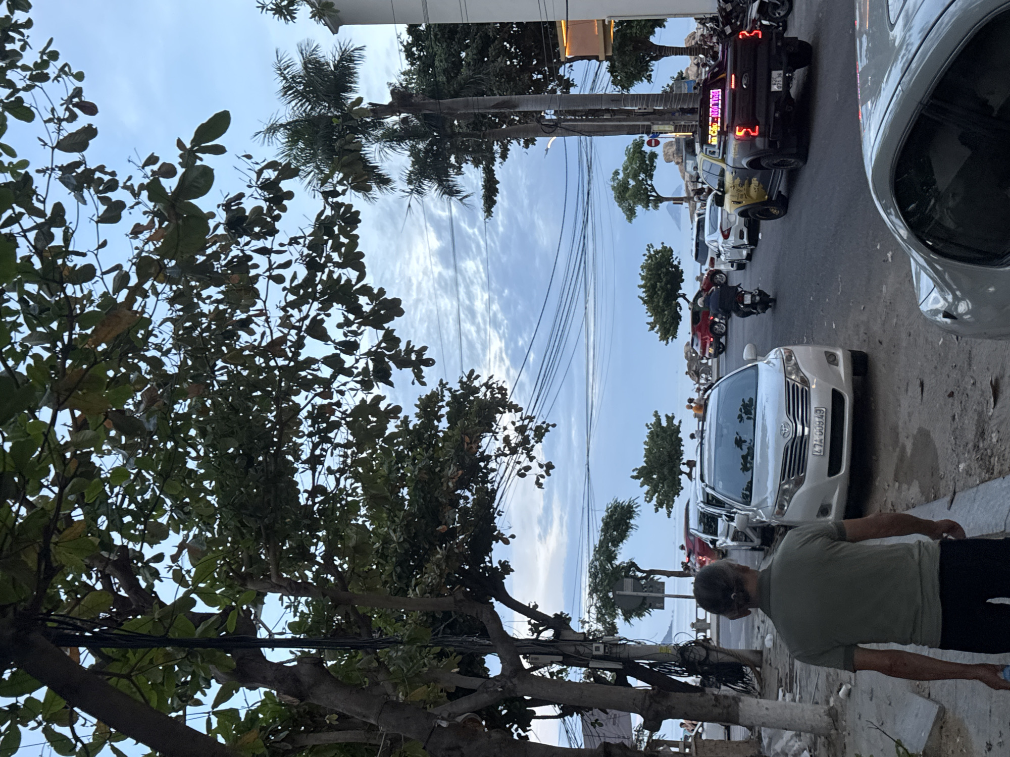 The image shows a street scene in Bac Nha Trang Ward, Vietnam. A man is walking along the sidewalk on the right side of the frame. The street is lined with parked cars and motorcycles. Tall trees are visible along the street and on the left of the frame, partially obscuring the sky and buildings. Utility wires are strung across the sky. In the background, the ocean and the beach can be seen.