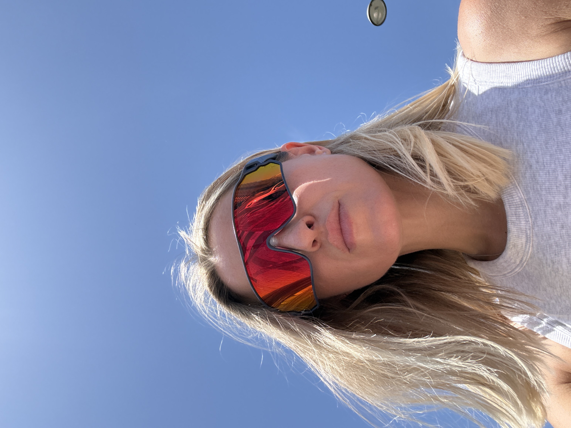 An extreme low-angle shot captures a close-up of a woman's face and upper chest against a clear blue sky. She is wearing large, wraparound sunglasses with mirrored red and orange lenses, reflecting the bright sunlight. Her blonde hair is windswept, framing her face. She has fair skin and appears to be looking upwards. Her lips are slightly parted. She is wearing a grey tank top. In the upper right quadrant of the image, a blurred street lamp is visible against the sky. The setting is outdoors, likely during the day, with the bright sky and lack of prominent shadows suggesting it is a sunny day. The absence of any distinguishable city landmarks or architecture makes it impossible to confirm the location as Valencia, Spain.