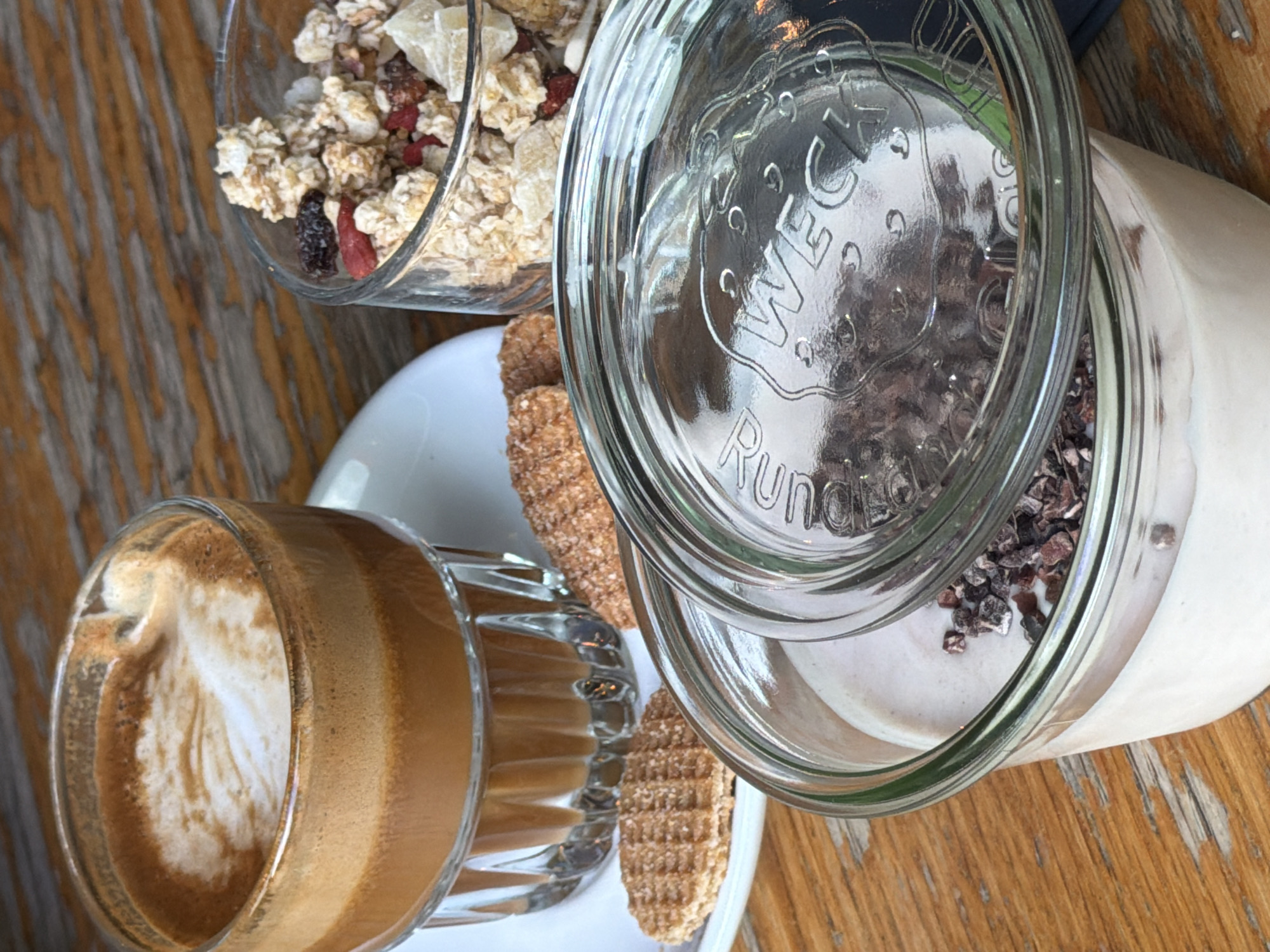 A close-up, eye-level shot shows a wooden table with a variety of breakfast items. On the left, a clear glass cup filled with coffee topped with foam sits on a white saucer. Next to the coffee cup are two waffle cookies and a small stack of three small, round cookies. To the right of the coffee, a clear glass jar filled with granola and dried fruit is positioned. In the foreground, a large clear glass jar with a silver lid lies on its side, partially covering a creamy substance topped with dark chocolate shavings. The lid is embossed with the word "WECK". The background is a blurred wooden surface. No people are visible. The scene does not contain any elements that would allow for specific identification of its location in Amsterdam, Netherlands.