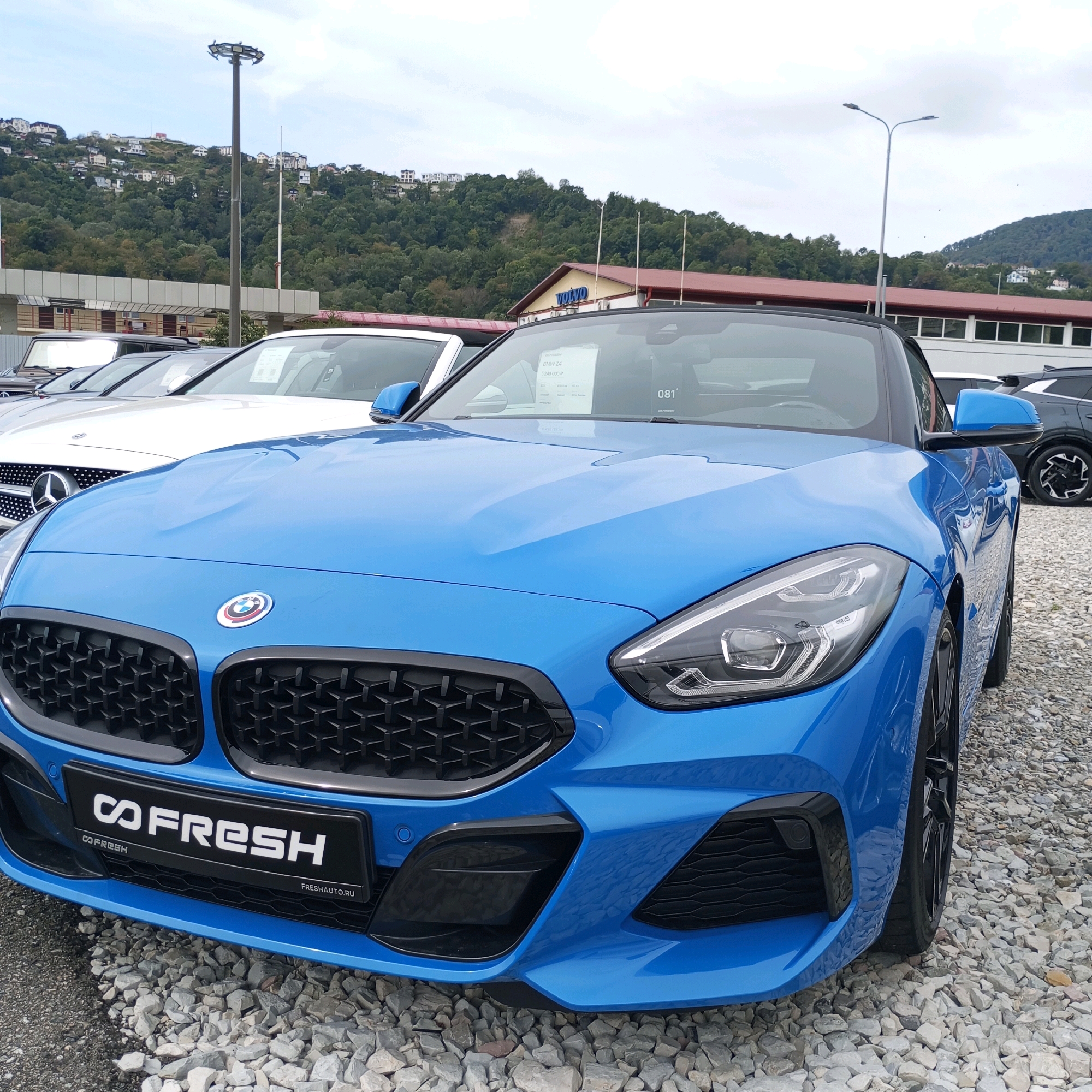 A bright blue BMW Z4 convertible is parked on a gravel lot in front of a building with a red roof. The building has "VOLVO" written on it in large blue letters. In the background, a lush green hillside is covered with houses and trees. The sky is overcast. The car is the primary subject, with its front end facing the viewer. The BMW logo is visible on the hood, and the license plate frame reads "CO FRESH." Other cars, including a white Mercedes-Benz, are parked behind the BMW. A tall light pole stands to the left of the frame. The scene appears to be a car dealership.