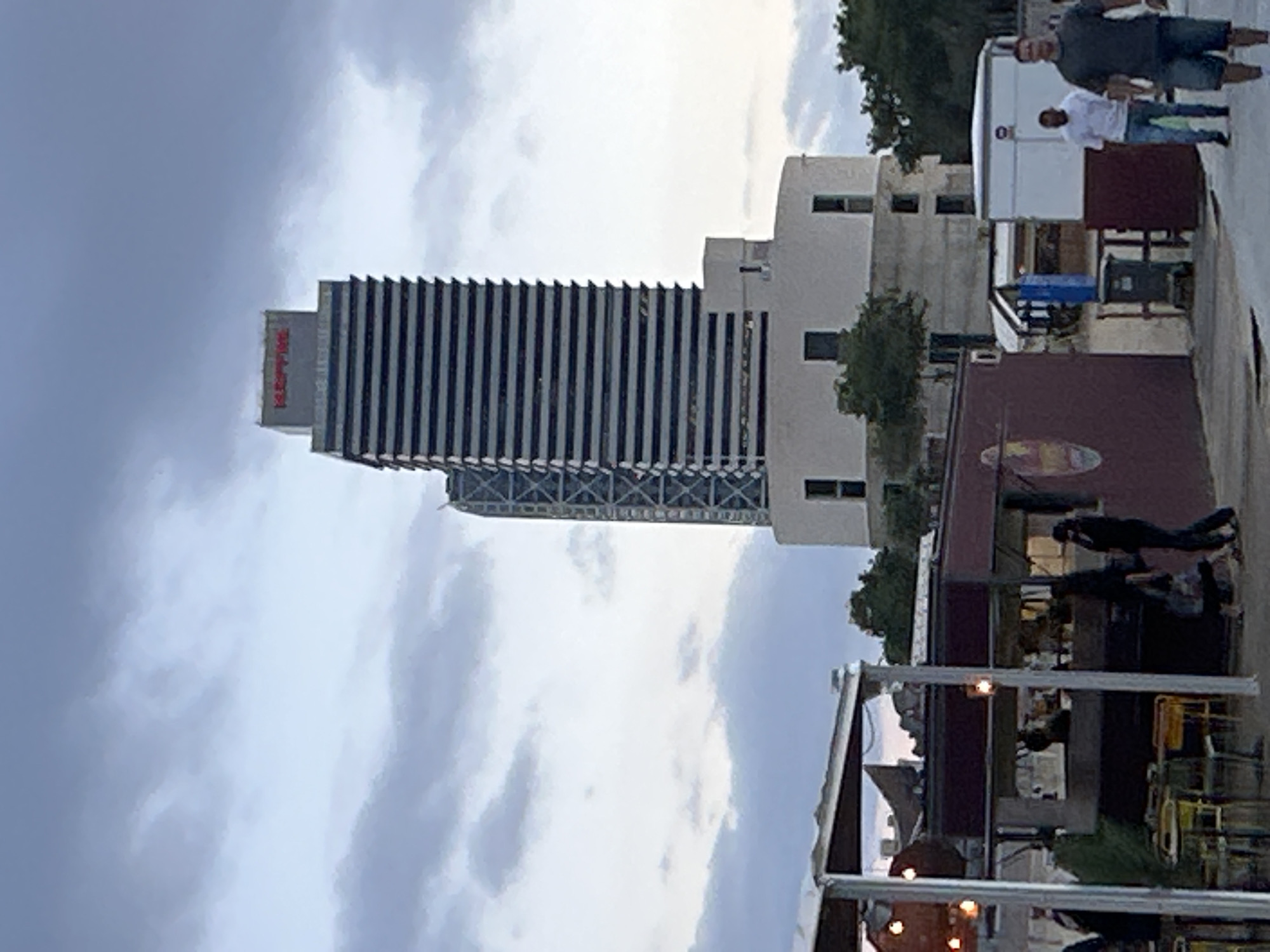 The image depicts the Hotel Arts in Barcelona, Spain, a prominent skyscraper located in the Port Olímpic district. The hotel, designed by Bruce Graham and Frank Gehry, is characterized by its distinctive glass facade and its striking height. In the foreground, a casual dining area with outdoor seating and food stalls is visible. People are seen walking and dining, enjoying the evening ambiance. The sky is overcast, with a mix of light and dark clouds, suggesting either dusk or an impending storm. The Hotel Arts is a well-known landmark in Barcelona, symbolizing the city's modern architectural achievements and vibrant urban life.