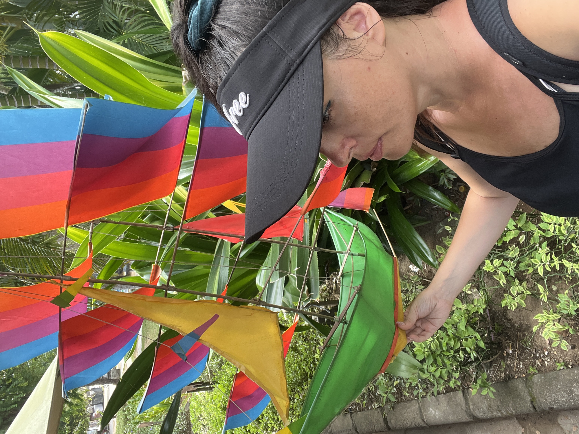 A woman wearing a black visor with the word "free" embroidered in white is shown in close-up, looking down at a collection of colorful kites. She is also wearing a black tank top. The kites are made of fabric and feature stripes of various colors, including blue, purple, red, orange, and yellow. Some kites are also green and yellow. The scene appears to be outdoors, with lush green foliage visible in the background. The lighting suggests it is daytime. The woman is gently touching one of the kites, possibly examining it or preparing it for flight. The overall mood seems relaxed and perhaps joyful, given the vibrant colors of the kites. The location context of Kuta Selatan, Indonesia, hints at a tropical environment.