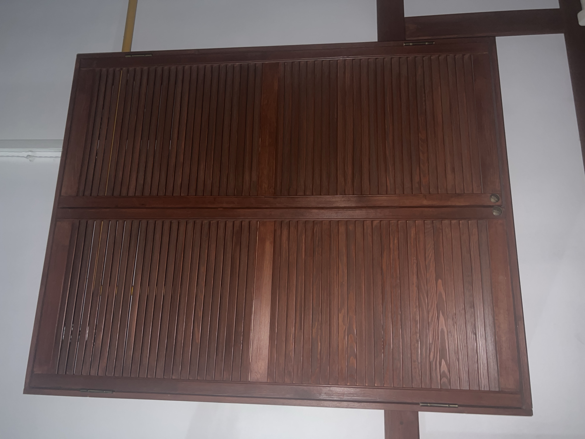 A pair of closed, dark brown louvered wooden shutters are mounted on a white wall. The shutters are divided down the center by a vertical seam and feature horizontal slats. Two small, circular knobs are visible at the bottom of the shutters, near the center. The shutters appear to be part of a window or a closet door. The lighting is from the right, casting a slight shadow on the left shutter.
