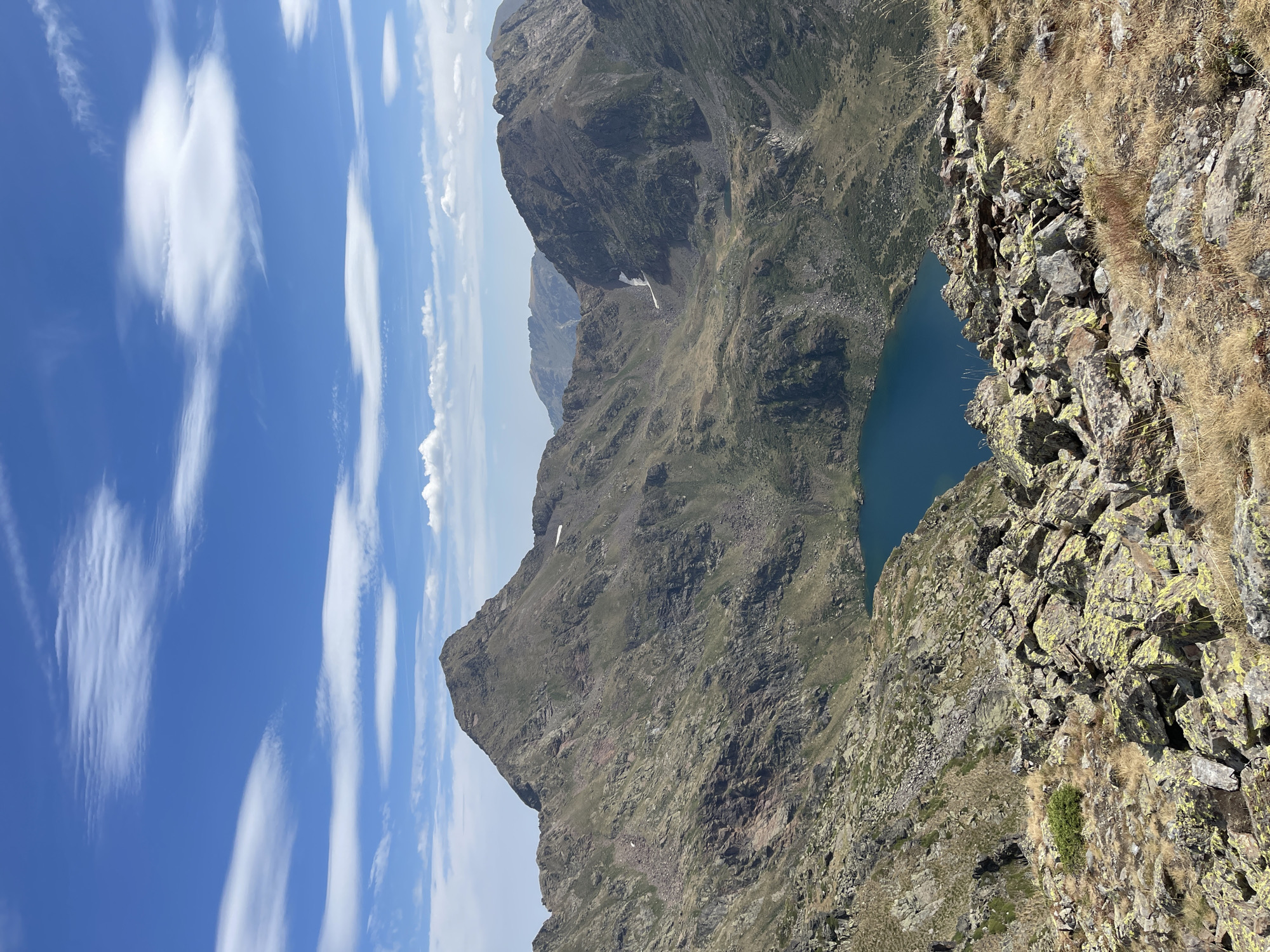 A high-angle, wide shot shows a vast, rocky mountain landscape under a bright blue sky with scattered white clouds. In the foreground, a rocky slope is covered with dry grass and lichen-covered rocks. Below this, a deep blue alpine lake is nestled in a valley. Jagged, barren mountain peaks dominate the middle ground and background, with patches of snow visible on some of the higher slopes. The overall impression is one of rugged, natural beauty in a remote, high-altitude environment.