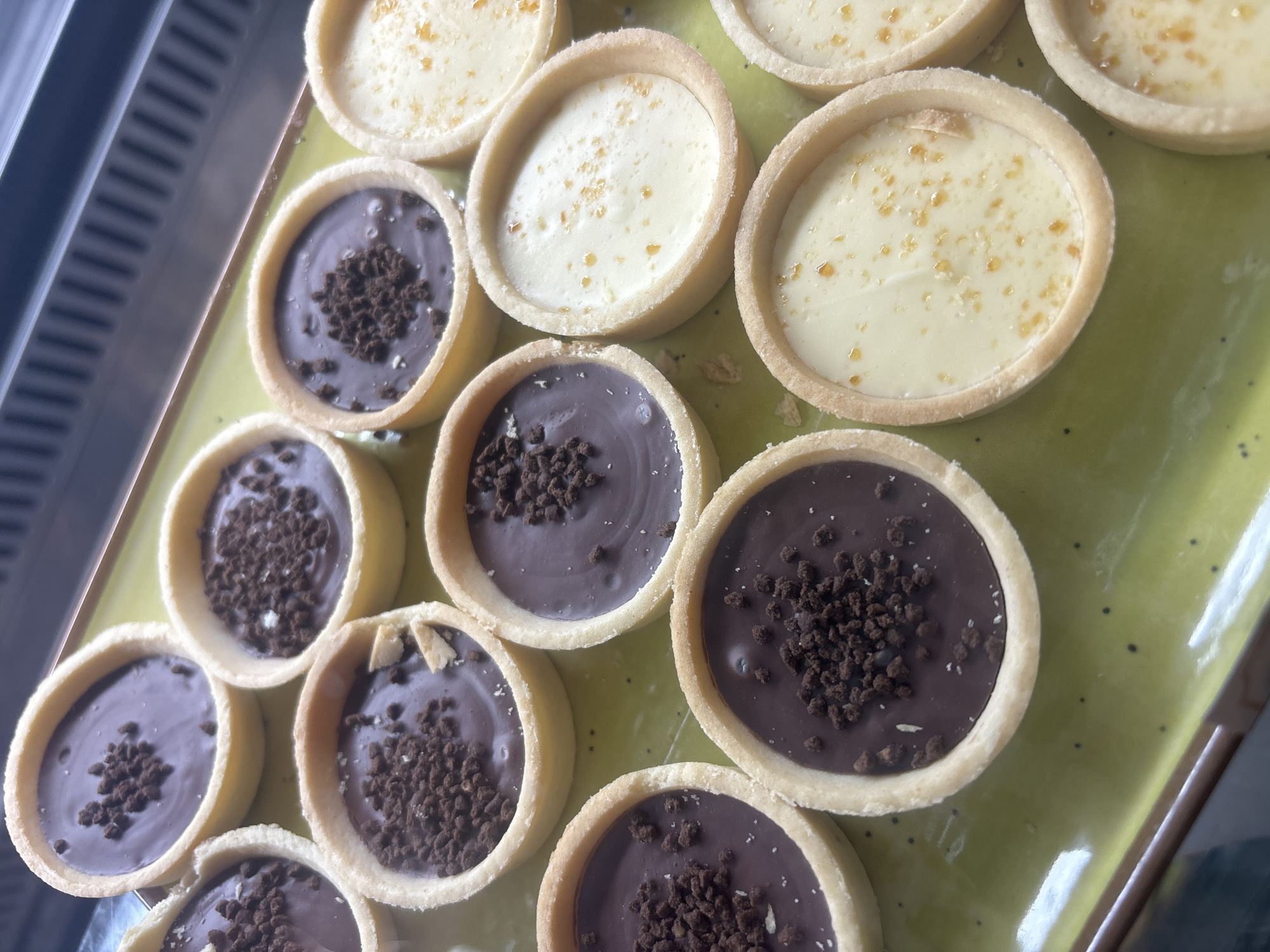 A top-down, slightly angled shot displays an assortment of mini tarts arranged on a light green, glossy tray. There are at least 15 tarts visible, neatly lined up in rows.
Approximately half of the tarts are filled with a creamy, light-colored (possibly vanilla or lemon) custard, topped with small, irregular orange or gold sprinkles, suggesting a candied zest or sugar crystal decoration. The other half are filled with a smooth, dark chocolate ganache, generously garnished with dark brown cookie crumbles or chocolate sprinkles.
The tart shells are a consistent light golden-brown, indicating a crisp, baked pastry. The tarts are uniform in size and round shape.
To the left of the tray, a blurred background shows vertical, reflective stripes, consistent with the shelves or glass of a refrigerated display case or bakery counter. This suggests the tarts are showcased for sale or ready to be served in an indoor setting, possibly a bakery, cafe, or a food display at an event. The overall impression is one of fresh, appealing pastries. No people or visible text are present in the image. The location context provided is Malki Iskar, Bulgaria.
