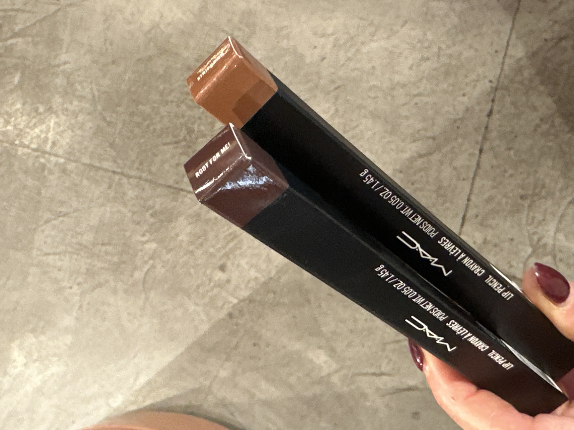 A close-up, high-angle shot captures a person's hand holding two MAC Lip Pencils in their black packaging against a light-colored, textured floor. The hand, adorned with dark red or burgundy nail polish, holds the two products horizontally.
Both product boxes clearly display the "MAC" logo in white, along with the text "LIP PENCIL CRAYON À LÈVRES" and the weight "POIDS NET WT. 0.05 OZ./1.45 g".
The ends of the lip pencils, protruding from their boxes, reveal their respective shades and names. The top pencil is a lighter, warm brown color, with partially visible text that appears to read "STRIPPED". The bottom pencil is a darker, rich brown shade, and its name is clearly visible as "ROOT FOR ME!".
The background is an indistinct, light gray or beige floor surface with subtle variations in tone and texture, possibly concrete or a large tiled area. The scene appears to be indoors. There are no specific visual cues within the image itself to confirm the provided location context of London, United Kingdom. The image focuses on the products and the act of holding them, possibly during shopping or product inspection.