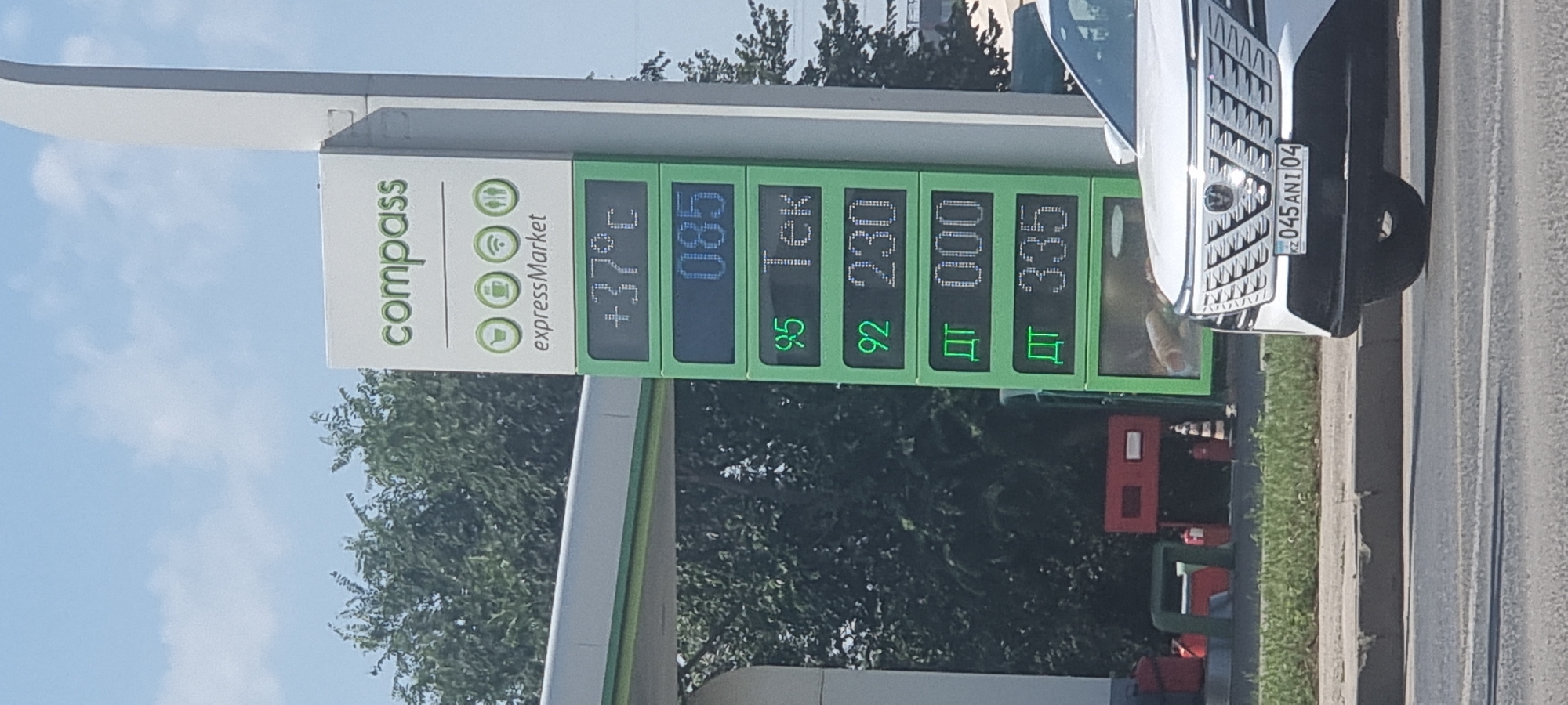 The image shows a street scene in Aktobe, Kazakhstan. A white car with license plate 045 ANI 04 is parked on the right side of the frame. In the background, a tall, green and white sign displays gas prices and the temperature. The temperature is shown as +37°C. Below the temperature, there are prices for different types of fuel, indicated by numbers like 95, 92, and quantities of fuel such as 000 and 335. The sign also has the words "compass" and "expressMarket". Trees and foliage are visible behind the sign. The sky is blue with some clouds.