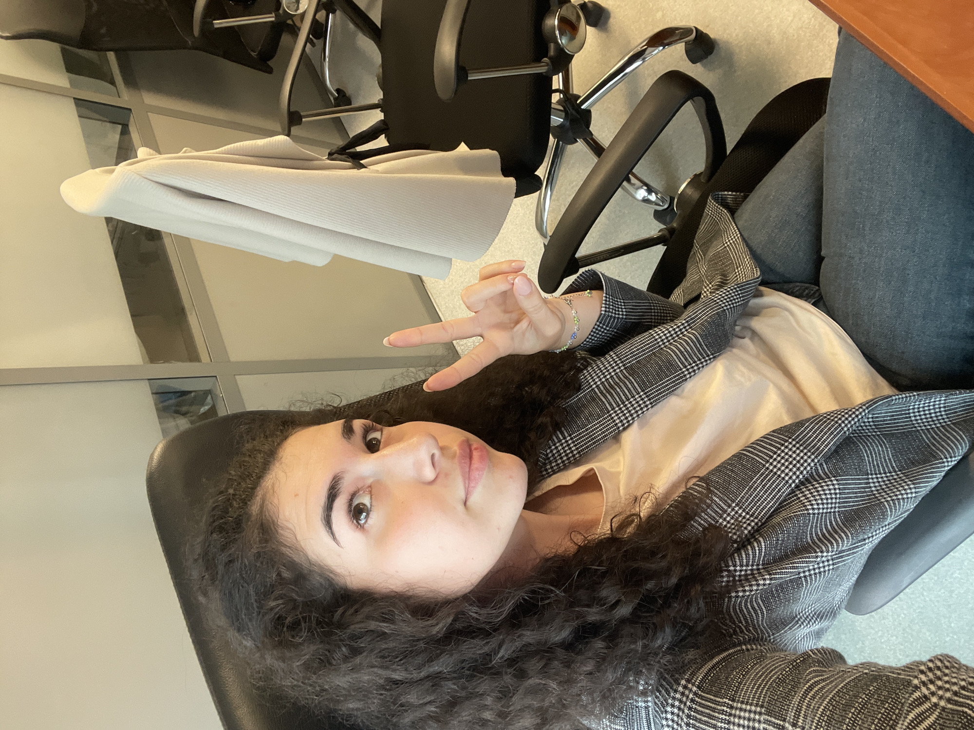 A selfie, taken in an office environment in Sofia, Bulgaria, features a young woman with long, dark, curly hair, seated in a black office chair. She is looking directly at the camera with a calm, neutral expression, and making a peace sign (V-sign) with her right hand, which is raised near her face. Her nails are manicured with a light polish, and a delicate, multi-colored beaded bracelet adorns her right wrist.
The woman is wearing a light-colored top, possibly cream or light pink, under a stylish grey and white plaid blazer. Her lower body, covered in dark blue jeans, is partially visible.
The background confirms an indoor office setting. Another black office chair with a chrome base is visible behind her, with a light-colored garment (perhaps a sweater or jacket) neatly folded and draped over it. To the right side of the frame, the edge of a wooden desk or table is present. The flooring appears to be a light-colored carpet or linoleum. In the distance, structures resembling glass office partitions or large windows can be seen, suggesting a modern workspace. The lighting throughout the image is bright and even, indicative of an interior environment, possibly during the daytime. There is no visible text in the image. The overall mood conveyed is relaxed and self-assured.