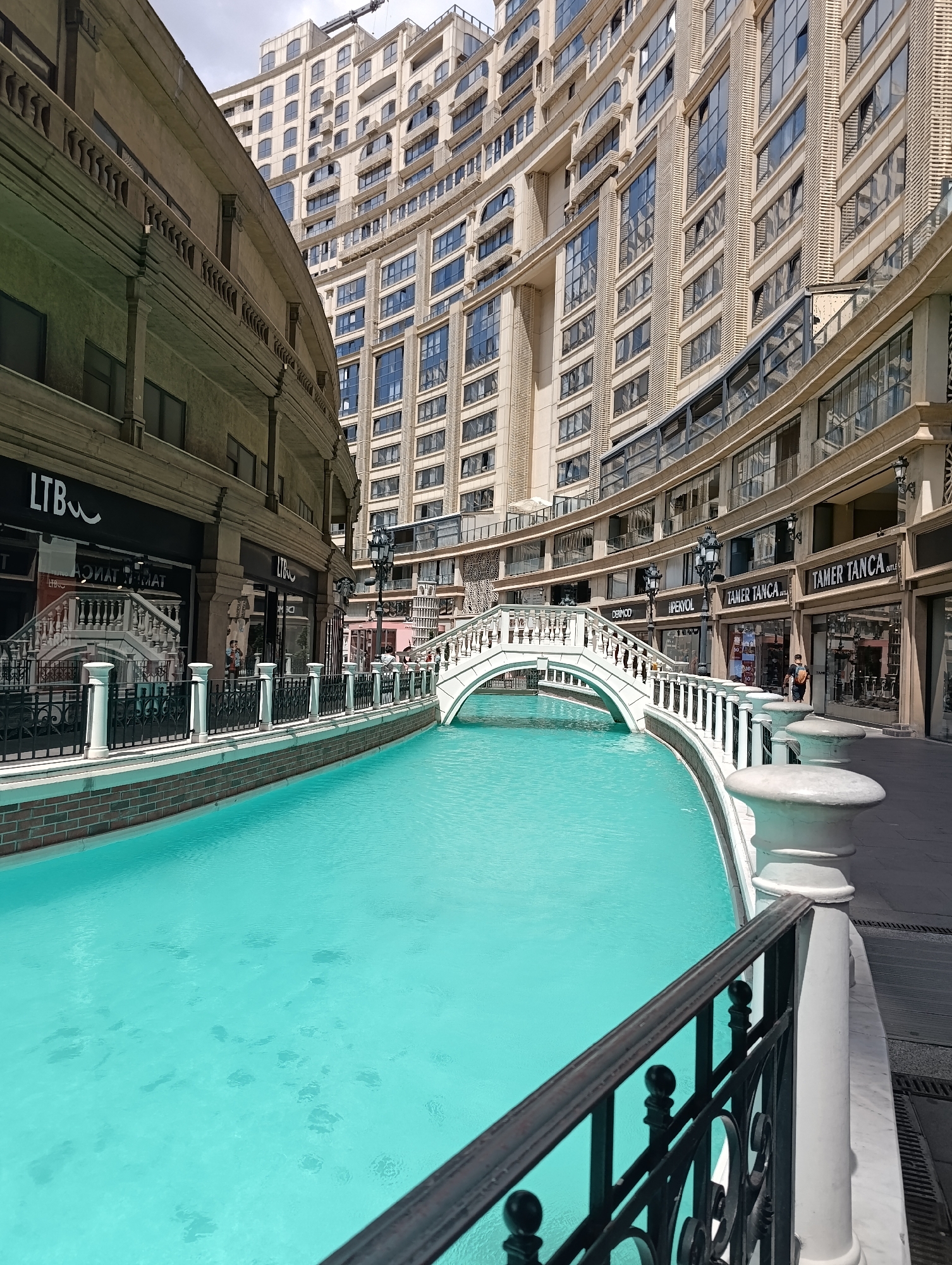 The image shows a view of the Venezia Mega Outlet in Gaziosmanpaşa, Turkey. In the foreground, a bright turquoise canal is visible, crossed by a white arched bridge. The canal is lined with a brick wall and a white railing with black metal accents. On the right side of the canal, a paved walkway runs alongside shops with storefronts displaying various brands. In the background, a tall, modern building with a curved facade and numerous windows dominates the skyline. The sky is overcast. Several people are present, walking along the shops and on the bridge.