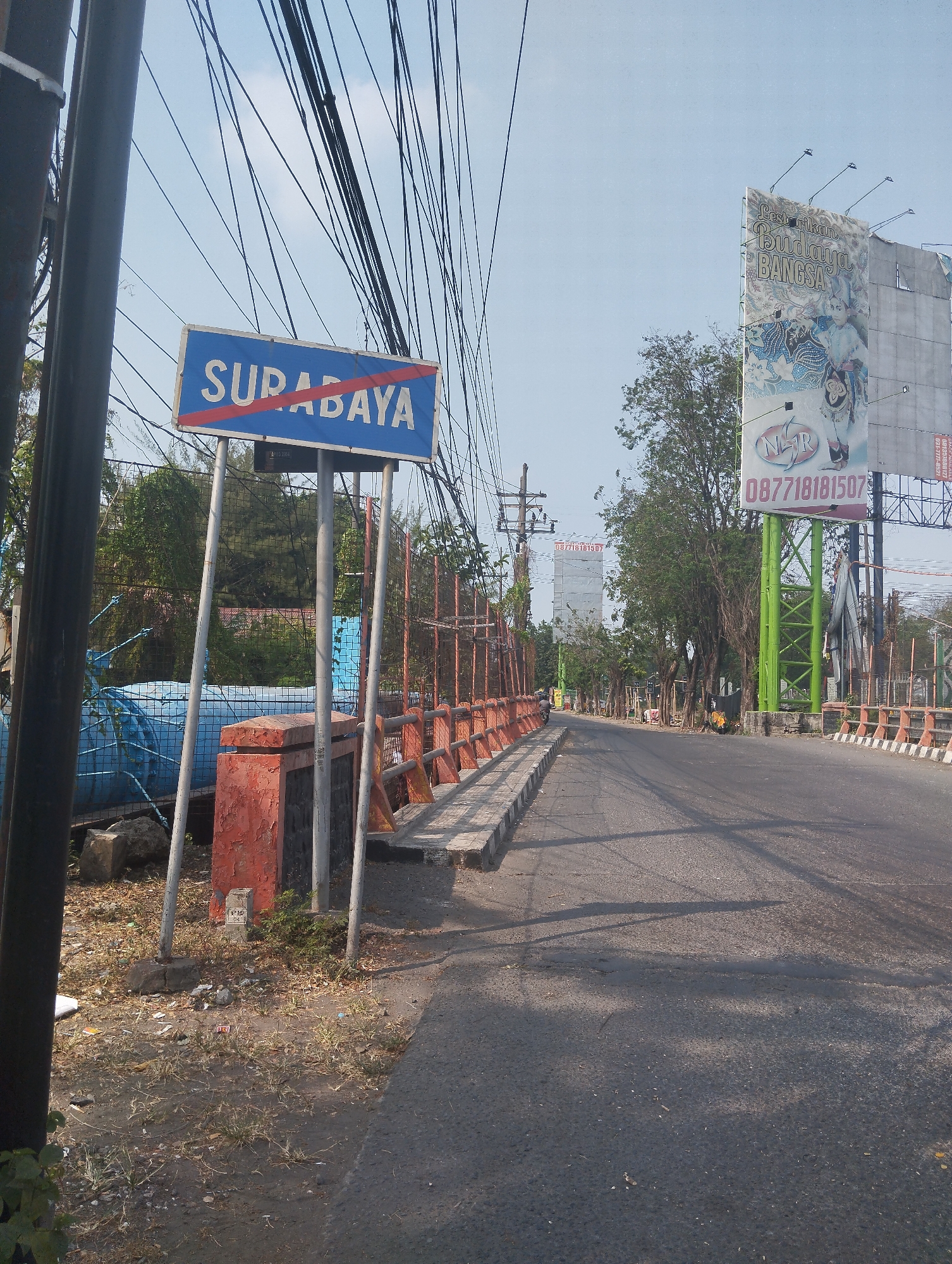 The image shows a road in Surabaya, Indonesia, with a sign indicating the city limits. The sign, which is blue with white lettering, reads "SURABAYA" and has red diagonal lines across it, suggesting it is a boundary marker or a prohibition. To the left of the sign, there is a large blue pipe and some vegetation. The road is paved and appears to be an urban area, with trees and buildings visible in the background. There are also numerous overhead wires, typical of power or telecommunication lines in the city. A billboard with text and images is visible on the right side of the road, advertising what appears to be a cultural event or product. The overall scene suggests a typical urban roadside environment in Surabaya.