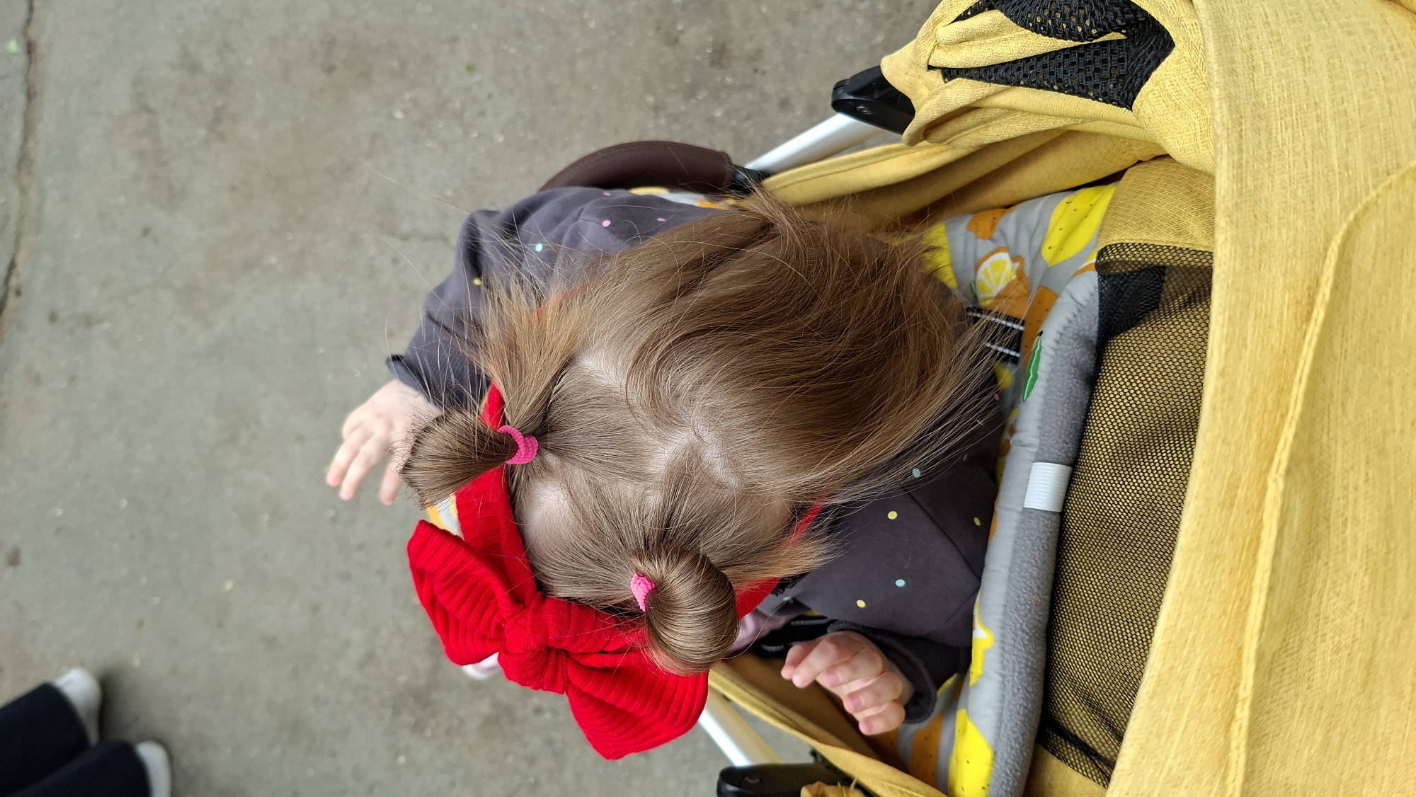 A top-down shot reveals a baby sitting in a yellow stroller on a concrete surface, possibly a sidewalk. The baby has light brown hair styled in two pigtails, each secured with a pink hair tie. A large red bow adorns one of the pigtails. The baby is wearing a dark gray shirt with multi-colored polka dots. The stroller has a yellow canopy, a gray and yellow patterned lining, and a black mesh storage area. A person's legs, clad in navy pants and white shoes, are partially visible in the lower left corner. The location is likely in an urban area, though not explicitly identified in the photograph.