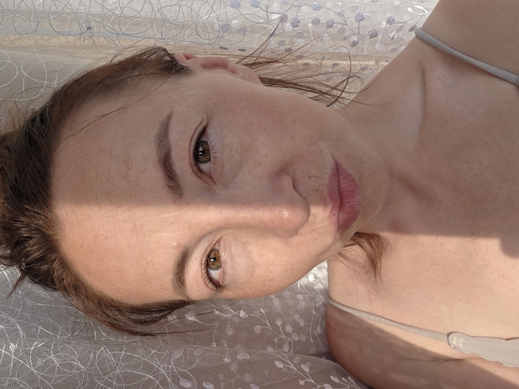 A close-up, head-and-shoulders shot of a woman with fair skin, brown hair, and brown eyes is captured against a white lace curtain in Chaikovsky, Russia. A diagonal shadow bisects her face from her forehead to her chin, highlighting the texture of her skin and the subtle freckles across her nose and cheeks. She is wearing a light-colored spaghetti strap top. The woman is looking directly at the camera with her lips slightly pursed, creating a playful expression. Her hair is pulled back loosely, with strands framing her face. The curtain behind her has a floral vine pattern. The lighting suggests it's daytime, with sunlight filtering through the curtain.