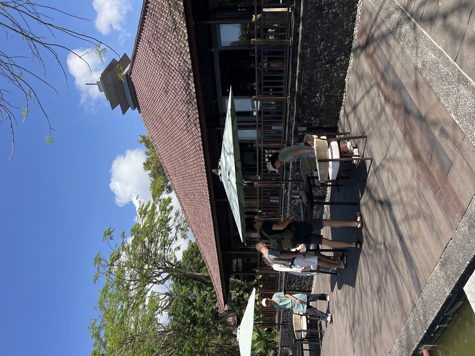 The image depicts an outdoor dining area of a restaurant in Baturiti, Indonesia, set against a backdrop of lush greenery and a clear blue sky with scattered clouds. A multi-tiered pagoda-like structure rises above the tiled roof of the restaurant building. Several tables with chairs are arranged on a wooden deck, some covered by large, light-colored umbrellas.
In the foreground, a person wearing a light green t-shirt and dark pants stands near a table, facing away from the camera and appearing to be in conversation with another individual. This second person, wearing a white cap and a light blue t-shirt, is also near a table.
Further to the right, a group of three individuals is clustered around a table. One person, with a backpack, is looking down at something on the table. Another person, wearing white shorts and a light-colored t-shirt, stands beside them. A third person, wearing a light green t-shirt and dark pants, is also near the table.
The wooden deck has a textured surface and is partially shaded by the surrounding trees. A stone wall borders the deck, and a small body of water is visible on the lower left side of the image. The restaurant building features large windows and a wooden railing. The overall atmosphere suggests a pleasant outdoor dining experience.