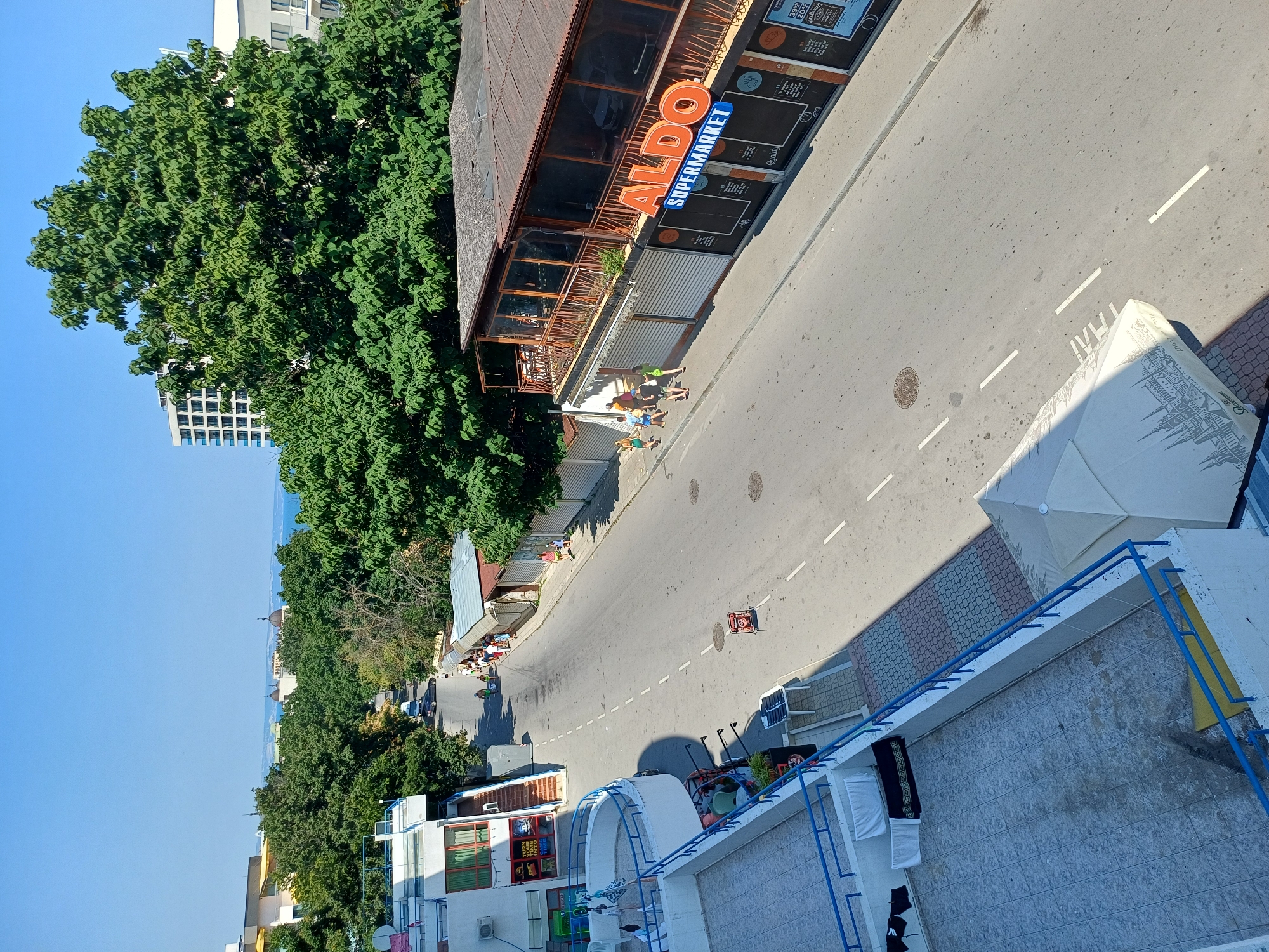 The image depicts a street scene in Varna, Bulgaria. In the foreground, a tiled rooftop with blue railings overlooks a paved street. Several items of laundry, including black t-shirts and white towels, are hung on the railing. To the right of the rooftop, a white umbrella is partially visible.
The street itself is asphalt with white dashed lane markings. Several people are walking on the sidewalk along the right side of the street. A building with a large orange sign that reads "ALDO SUPERMARKET" is prominent on the right. This building has a covered outdoor seating area with wooden railings and glass enclosures. Other buildings line the street, including what appears to be a multi-story white building with balconies on the left. Trees with lush green foliage are scattered throughout the scene, some obscuring views of taller buildings in the background. The sky is clear and blue.
Further down the street, several more people are visible, and a few vehicles are parked or driving. In the distance, a glimpse of the sea and more buildings can be seen. The overall impression is a sunny day in a coastal town with a mix of commercial establishments and residential buildings.