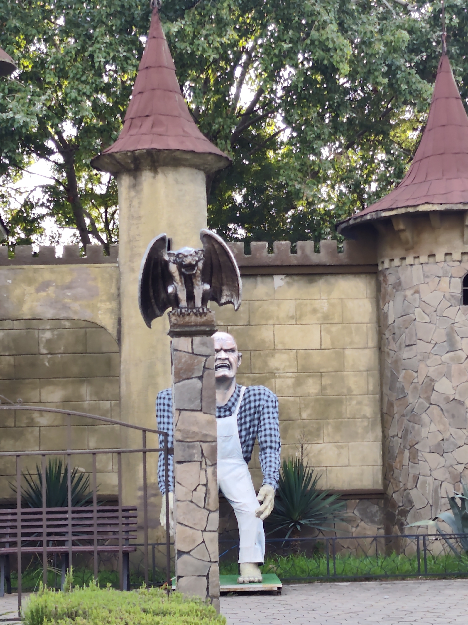 This image depicts a peculiar outdoor scene, likely a themed attraction, featuring a castle-like structure and two unusual figures. The castle wall is constructed from large, irregular stones on the right side, transitioning to a lighter-colored, brick-patterned surface on the left. Two conical, reddish-brown turrets rise above the wall, one to the left and another to the right, each topped with a finial. Lush green foliage from trees with dense leaves forms a canopy overhead, with sunlight filtering through.
In the foreground, a dark metal fence encloses a garden area. To the left of the fence, a dark brown bench with a slatted seat and back is visible. A spiky, dark green plant is positioned behind the bench.
Dominating the central portion of the image are two figures. The first, a gargoyle statue, is perched atop a stone pillar to the left. It is dark gray, with outstretched wings and a monstrous face. Partially obscured by the gargoyle's pillar is a large, humanoid figure. This figure has a bald, pale head with a contorted, menacing expression, revealing teeth. It wears a blue and black checkered shirt and white overalls. The figure appears to be standing on a wooden platform. The overall aesthetic suggests a theme park or a decorative installation, possibly related to fantasy or horror elements.
The location is identified as Sochi, Russia. The scene is set outdoors, with paved ground in the foreground, suggesting a public area or walkway. The lighting indicates it is daytime.