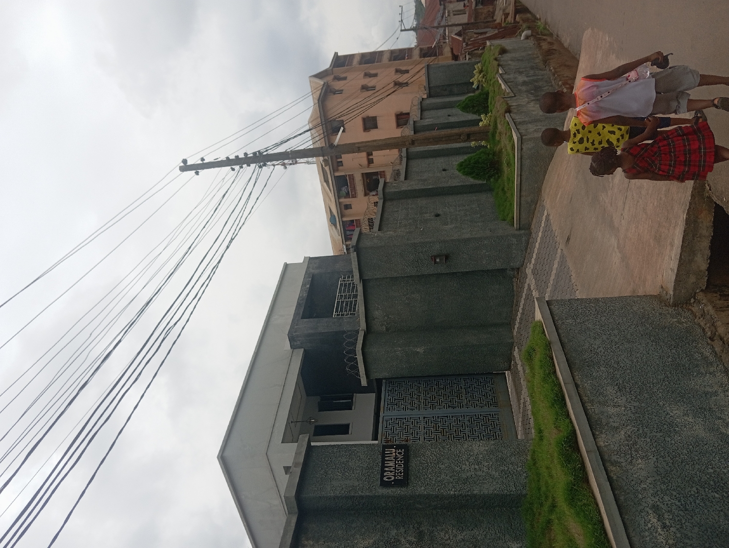 This image, taken in Nkpor, Nigeria, depicts an urban street scene under an overcast sky during the daytime. The perspective is slightly low, looking up towards a dense network of overhead utility wires connected to a tall pole, upon which several birds are perched.
In the foreground, a modern residence, identified by a plaque as "ORAMALU RESIDENCE," is prominent on the left. The building features dark grey and black textured walls and an intricately patterned metal gate at its entrance. Lush green grass landscaping runs along the front of the property.
On the right side of the frame, three children are walking away from the camera along a concrete sidewalk. The child furthest from the viewer wears a white top over an orange undershirt and shorts. The middle child is dressed in a yellow patterned shirt and dark shorts. The closest child, a girl, wears a red and black plaid skirt and a dark top.
In the background, behind the foreground residence, a multi-story building with a light brown/tan facade is visible. The numerous utility wires crisscrossing the sky are a notable feature, indicating a typical urban or semi-urban environment. The image captures a moment of daily life on a residential street.