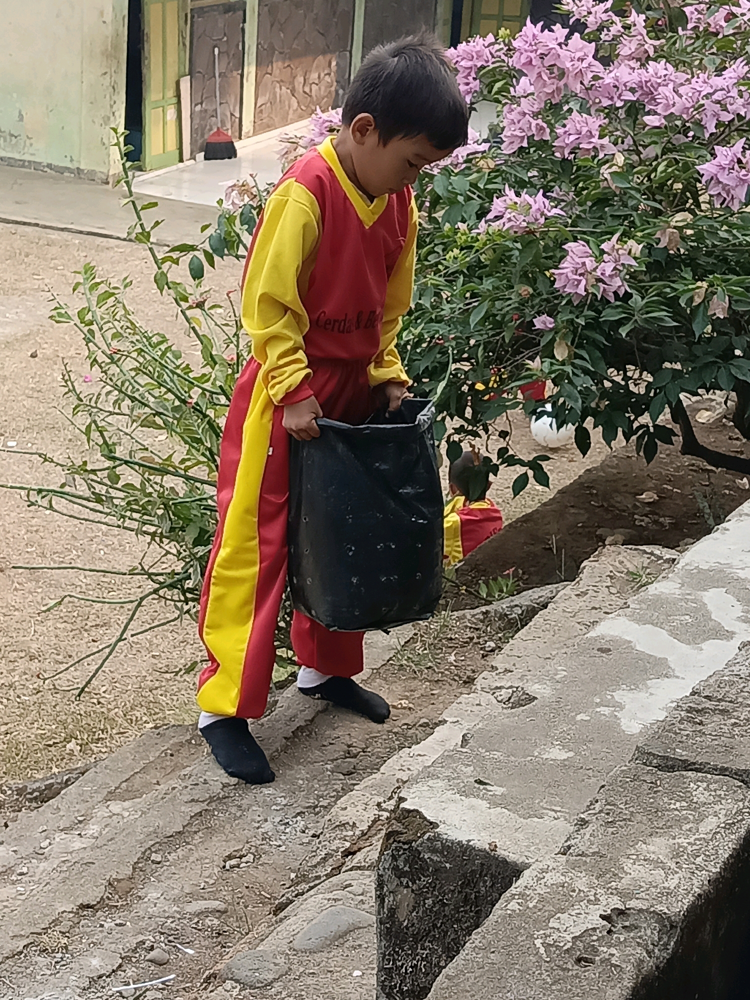 A young boy wearing a red and yellow uniform stands on a concrete step, looking down at a large black trash bag he is holding. He is outdoors, near a building with green and brown walls and a yellow door. A bush with pink flowers is to his right. The ground is covered in gravel and some dry vegetation. The scene appears to be in an outdoor area, possibly a school or community space in Taba Tinggi, Indonesia.
