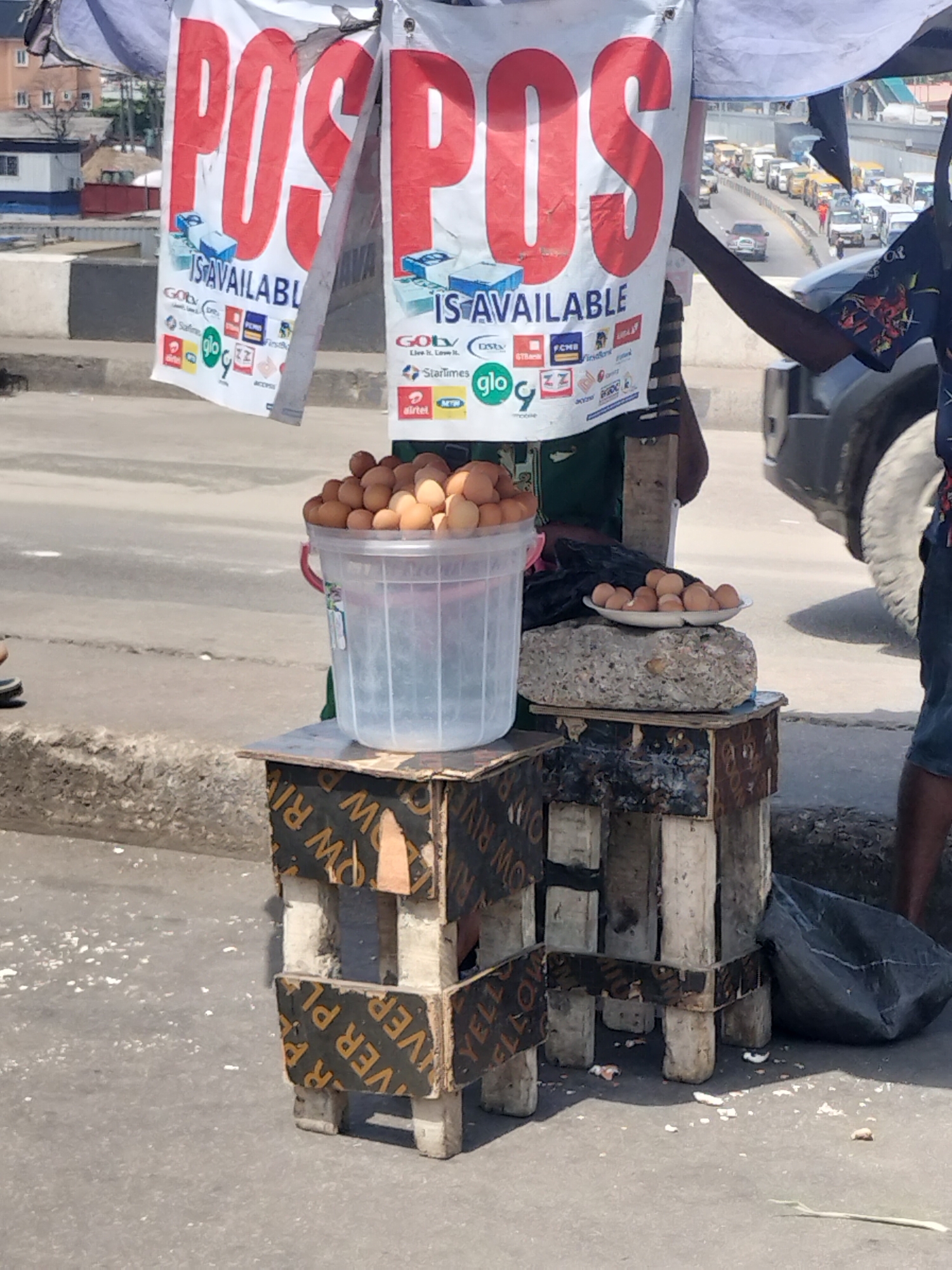 This image captures a street vending scene likely set in Lagos, Nigeria, under bright daylight, possibly near an overpass or busy urban thoroughfare. The weather appears sunny with distinct shadows cast.
In the foreground, a makeshift stall is constructed from repurposed wooden planks and plywood, some pieces bearing the inverted text "YELLOW RIVER PLYWOOD." On the left side of the stall, a large clear plastic bucket is filled to the brim with numerous brown eggs. On the right, a smaller quantity of eggs rests in a white dish, placed on a rough concrete block.
Above the stall, two white banners are prominently displayed, featuring large red text that reads "POS POS IS AVAILABLE." Below this, a collection of logos from various Nigerian telecommunications and financial service providers is visible. These include GOtv, DStv, StarTimes, Airtel, MTN, Glo, FCMB, FirstBank, GTBank, Zenith Bank, and Access Bank, indicating that mobile payment services are offered at this street-side setup.
A person with dark skin, wearing shorts and a patterned shirt, is partially visible on the right, standing next to the stall. Their outstretched arm suggests they might be tending to the goods or interacting with a customer. Another person's feet are barely visible on the far left.
In the background, a busy road or highway with significant vehicle traffic stretches into the distance, reinforcing the urban setting. The scene portrays a common sight of informal commerce, blending traditional hawking with modern payment solutions, typical of bustling Nigerian cities. A black plastic bag is on the ground near the right stand.
