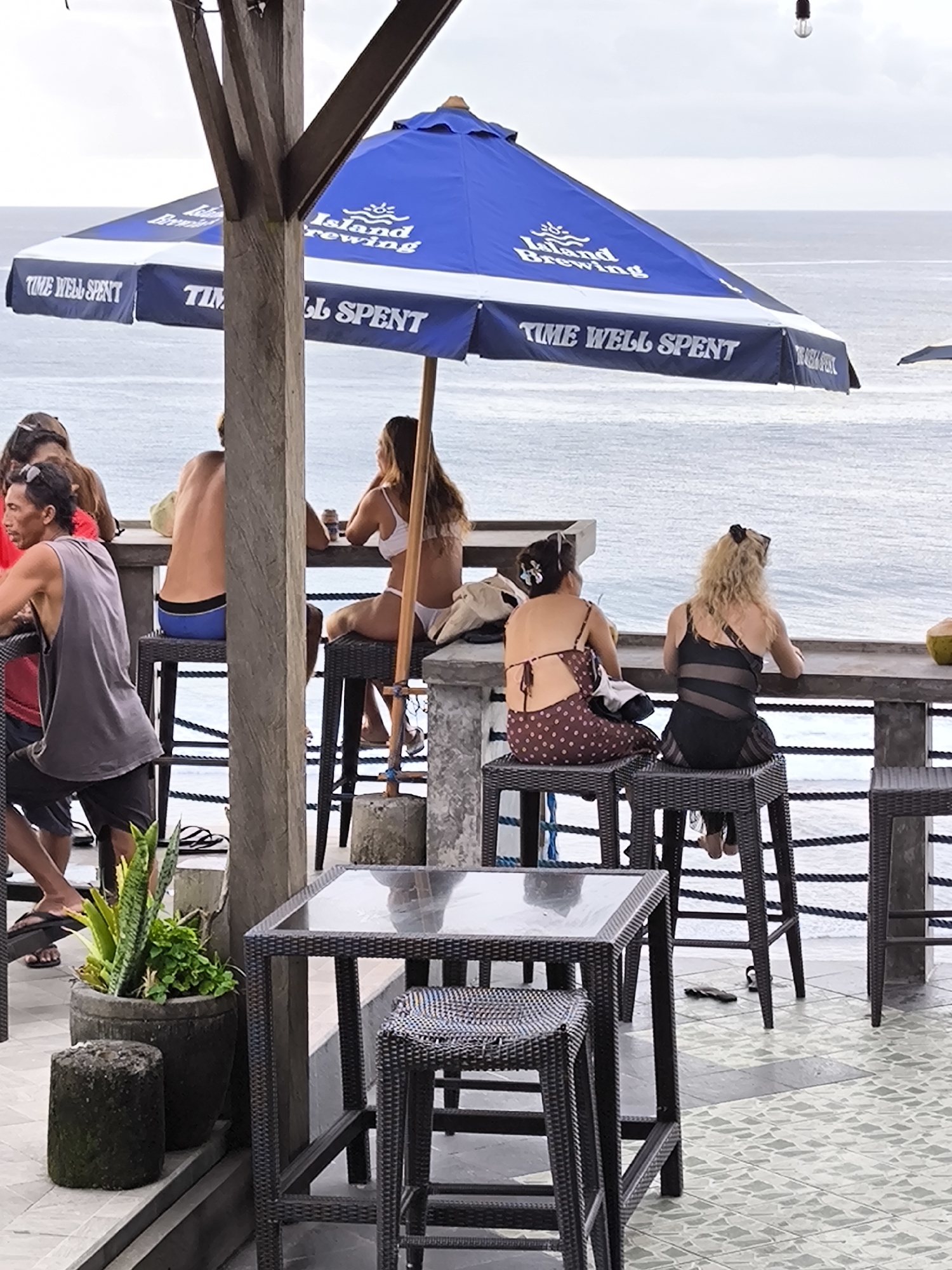 The image captures a relaxed scene in Uluwatu, Indonesia, likely at a beachfront bar or restaurant. Several people are seated on high stools at a bar counter, facing the ocean. A large blue and white umbrella, branded with "Island Brewing" and the phrase "Time Well Spent," provides shade. The foreground features a dark wicker table with a small stool, and a potted plant sits on the left. The ocean serves as the backdrop, with a slightly overcast sky. The scene suggests a casual, tropical ambiance.