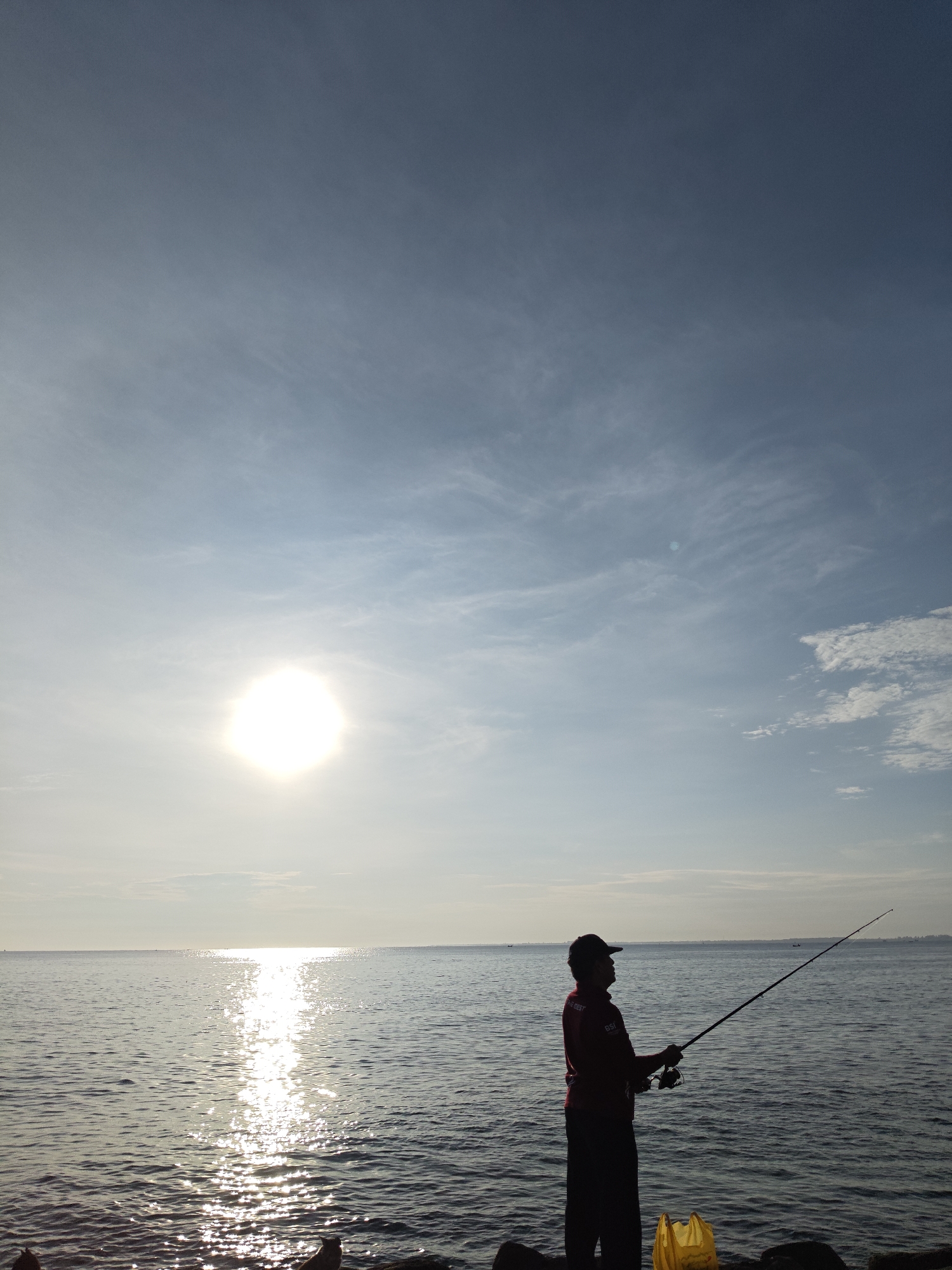 A silhouetted man stands on the shore of a body of water, holding a fishing rod. The sun is low in the sky, creating a bright reflection on the water's surface. A plastic bag sits at his feet. The location is likely Lhokseumawe, Indonesia.