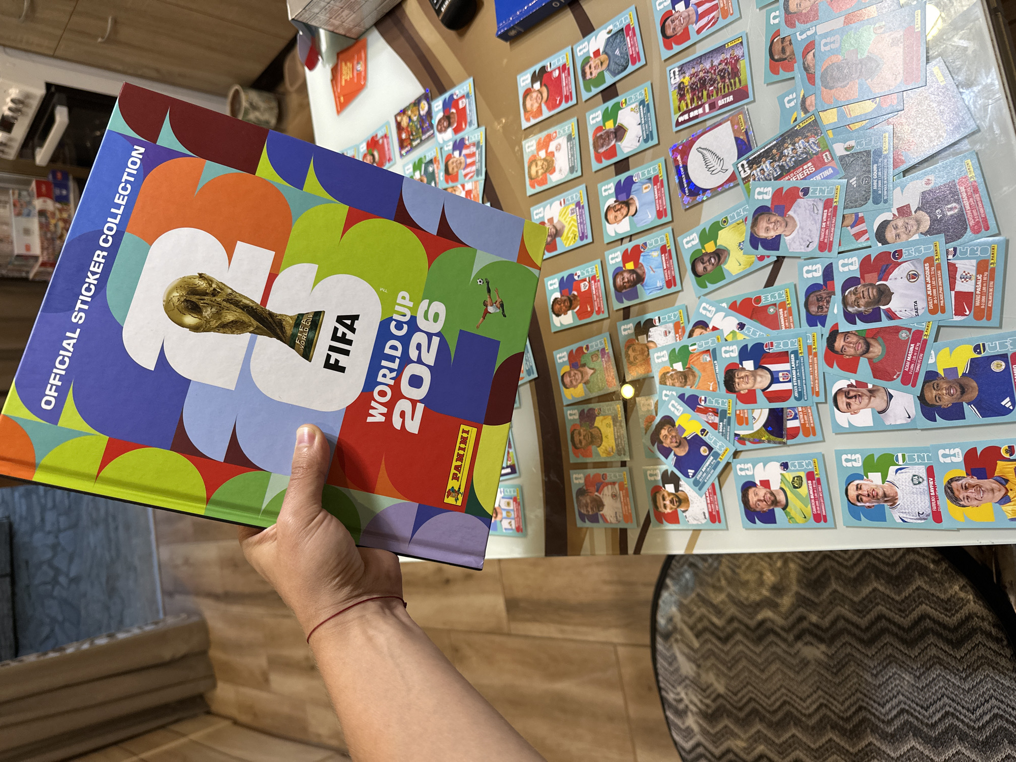 In the city of Prisovo, Bulgaria, a person's hand holds up a "FIFA World Cup 2026 Official Sticker Collection" book. The book's cover features a colorful design with the FIFA World Cup trophy and the text "FIFA WORLD CUP 2026". Scattered on a table below the book are numerous loose FIFA World Cup 2026 trading cards featuring images of soccer players. The background shows a wooden floor and a partially visible piece of furniture.