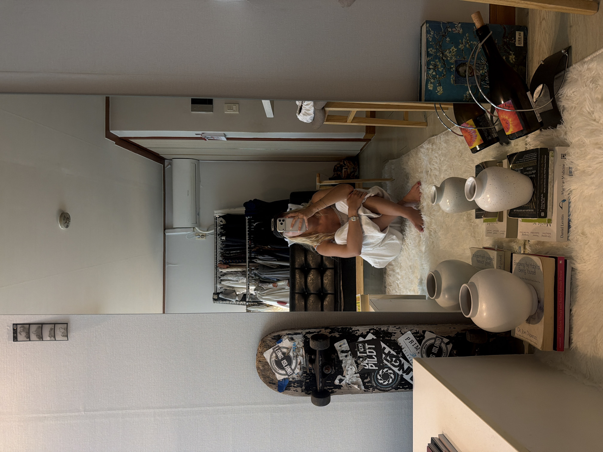 The image depicts a mirror reflection of a room interior. A person, presumably female, is seen holding a smartphone, seemingly taking a picture in the mirror. She is partially covered with a white towel or sheet. The reflection also shows a closet with various clothing items.
In the foreground, objects are arranged on a surface including: wine bottles in a metal holder, several books stacked next to each other, white decorative vases. A skateboard with stickers is visible.
The environment is indoor, and the room's decor includes a fluffy white rug.
There is no verifiable information to determine this to be in Seoul, South Korea.
