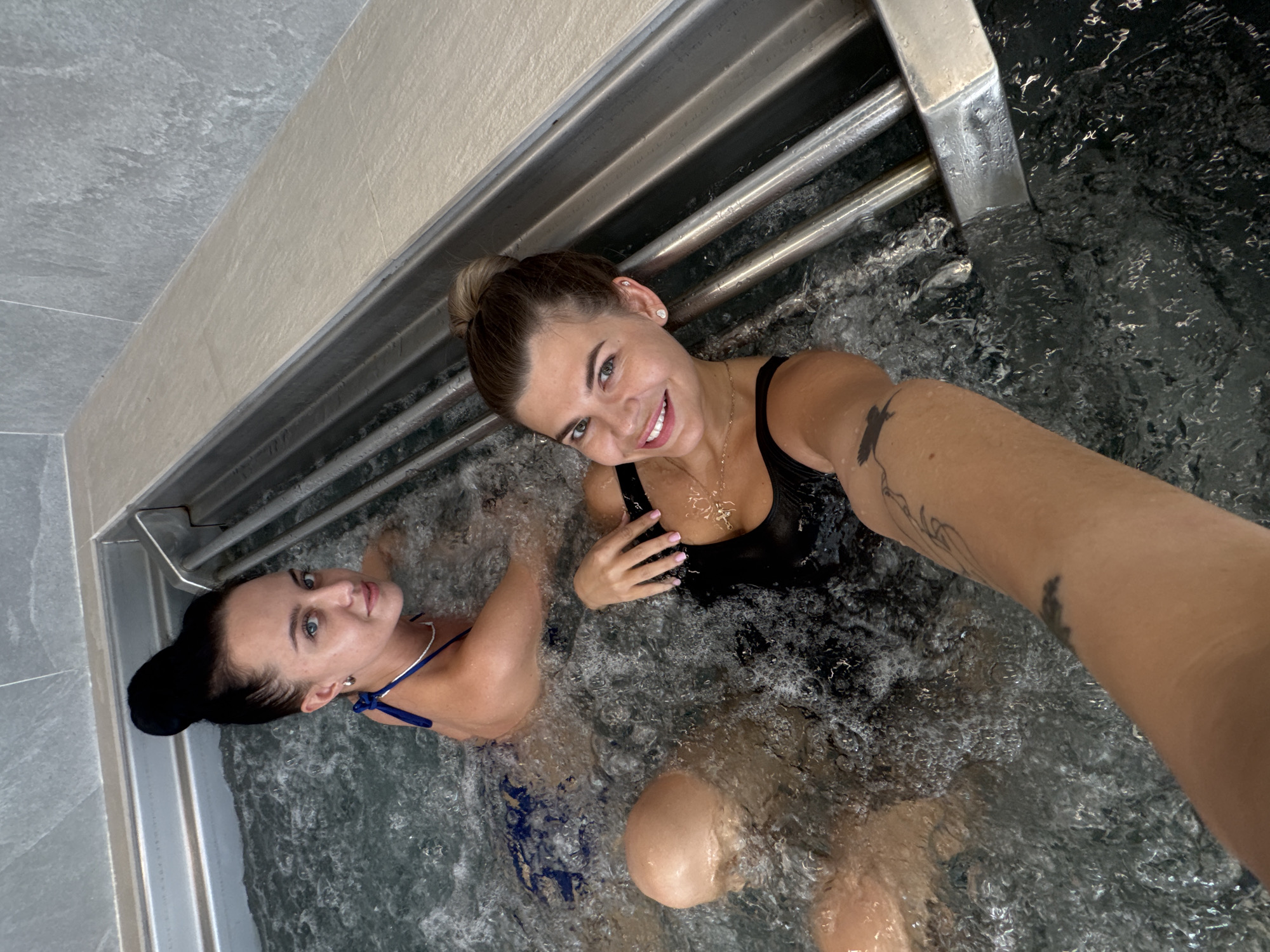 Two women are in a hot tub filled with bubbling water. One woman has her hair in a bun and is wearing a black swimsuit and necklace. The other woman has dark hair pulled back and is wearing a blue swimsuit. The hot tub is surrounded by gray tiles and a metal railing. The image is taken from a high-angle perspective. It appears to be a selfie. There is no information available in the image to confirm the location in Chaikovsky, Russia.