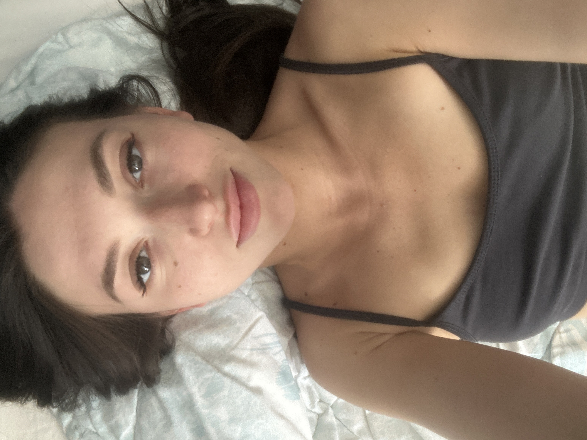 A young woman with dark brown hair is lying on a light blue and white patterned comforter. She is looking directly at the camera, with her head tilted slightly back. She has large, expressive brown eyes with black eyeliner and mascara. Her eyebrows are dark and arched. Her nose is small and slightly upturned. Her lips are full and have a subtle pink lipstick. She is wearing a dark grey tank top, with thin straps. Her skin is fair and has a scattering of light freckles across her cheeks and chest. The overall lighting is soft and natural, suggesting an indoor setting. The image appears to be a selfie. There is no discernible information about the city of Эстачи, Russia in this image.