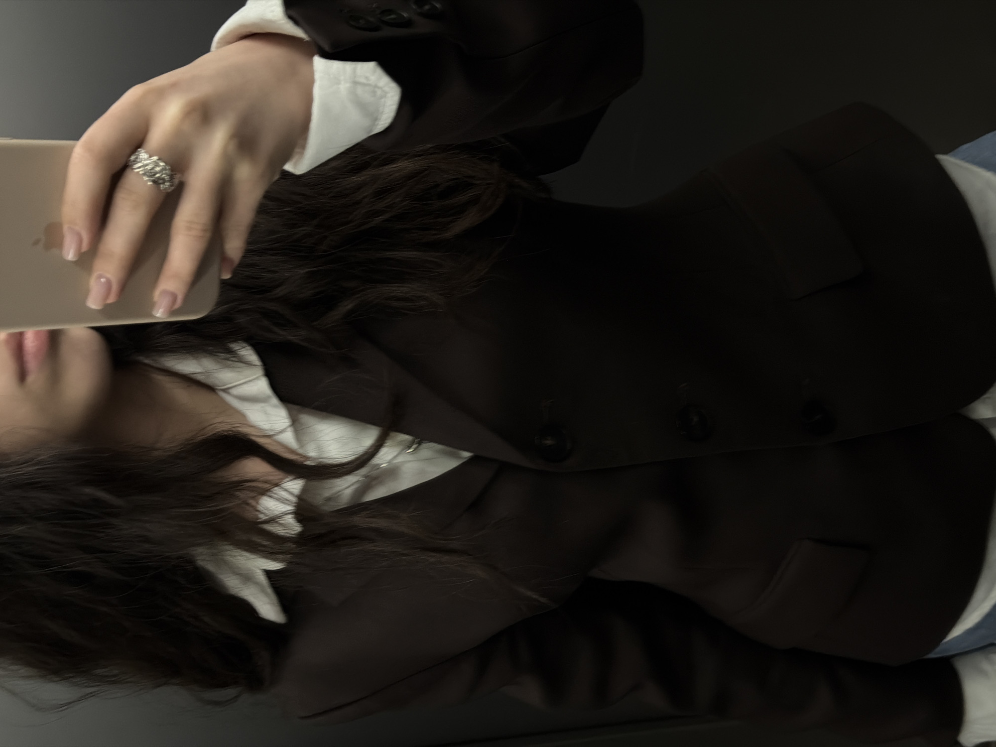 A woman wearing a dark blazer over a white collared shirt is holding a smartphone in her left hand, which is adorned with a silver ring. The scene takes place indoors, indicated by the ambient lighting and absence of exterior elements. The location is not discernible as Yerevan, Armenia, from the provided visual context.