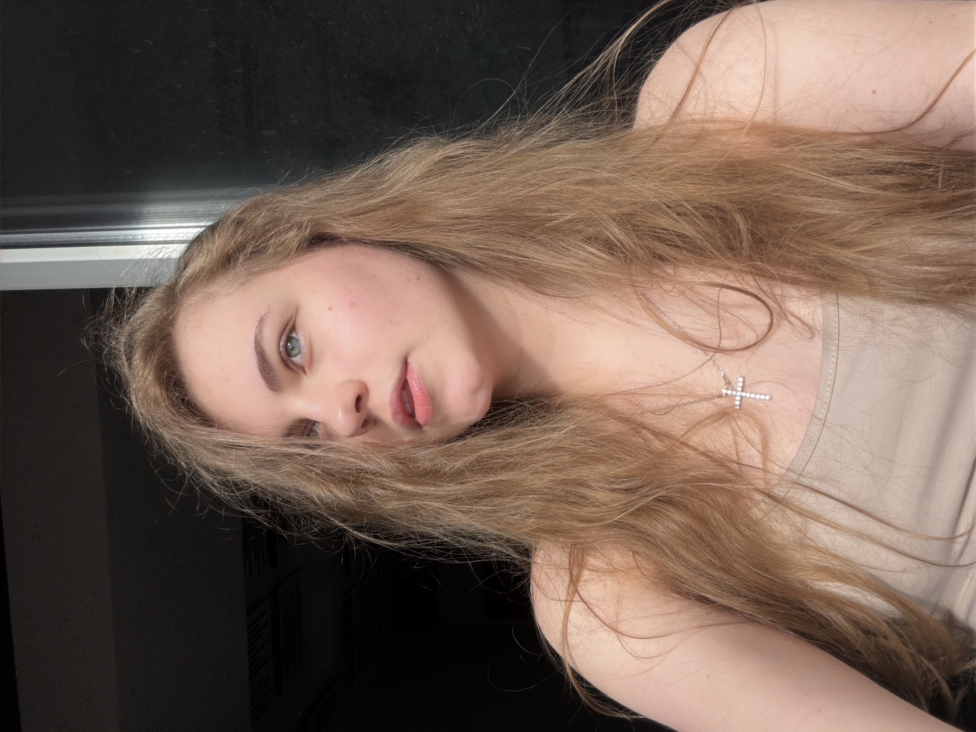 A young woman with long, blonde, wavy hair is shown from the chest up. She is looking to her left, with her head tilted slightly. She has fair skin, blue-green eyes, and pink lips. She is wearing a light-colored tank top and a silver cross necklace. The background is dark, with a bright vertical light source to her right, possibly a window or a door. The image was taken indoors. The location is not specified in the image or metadata.