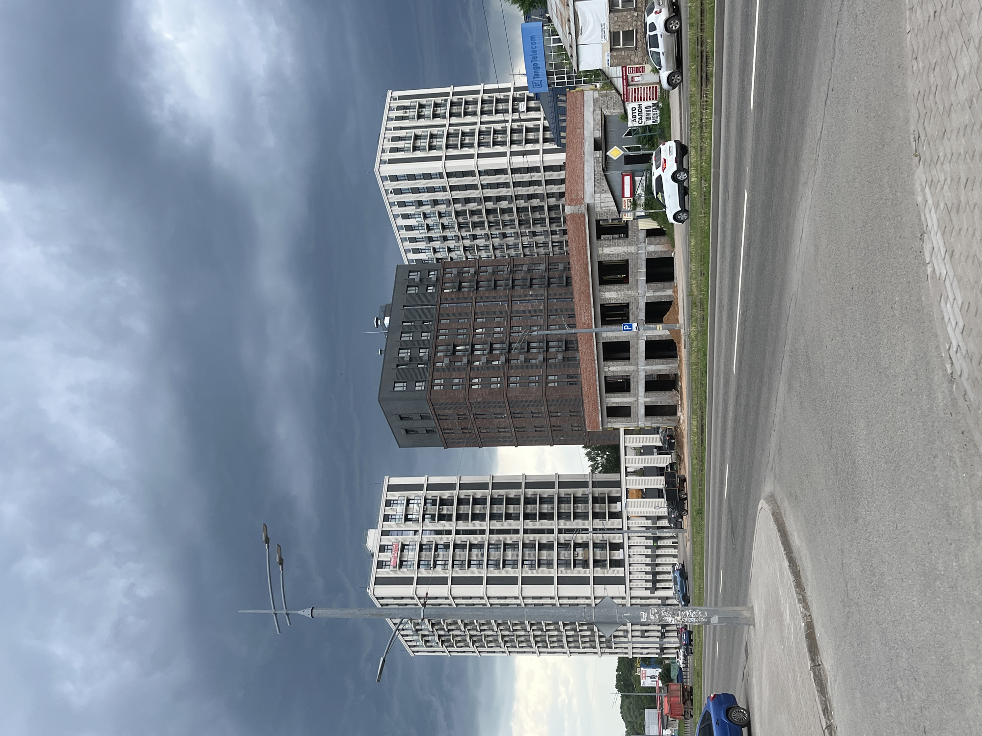 The image shows a street scene in Izhevsk, Russia, with a focus on several buildings. Three high-rise buildings are prominently displayed, including one under construction with visible brickwork. The other two appear to be residential apartments or office buildings. The sky is overcast, indicating potentially inclement weather. A street with white lane markers runs parallel to the buildings. A lamp post stands in the foreground. Several vehicles are parked along the street.
