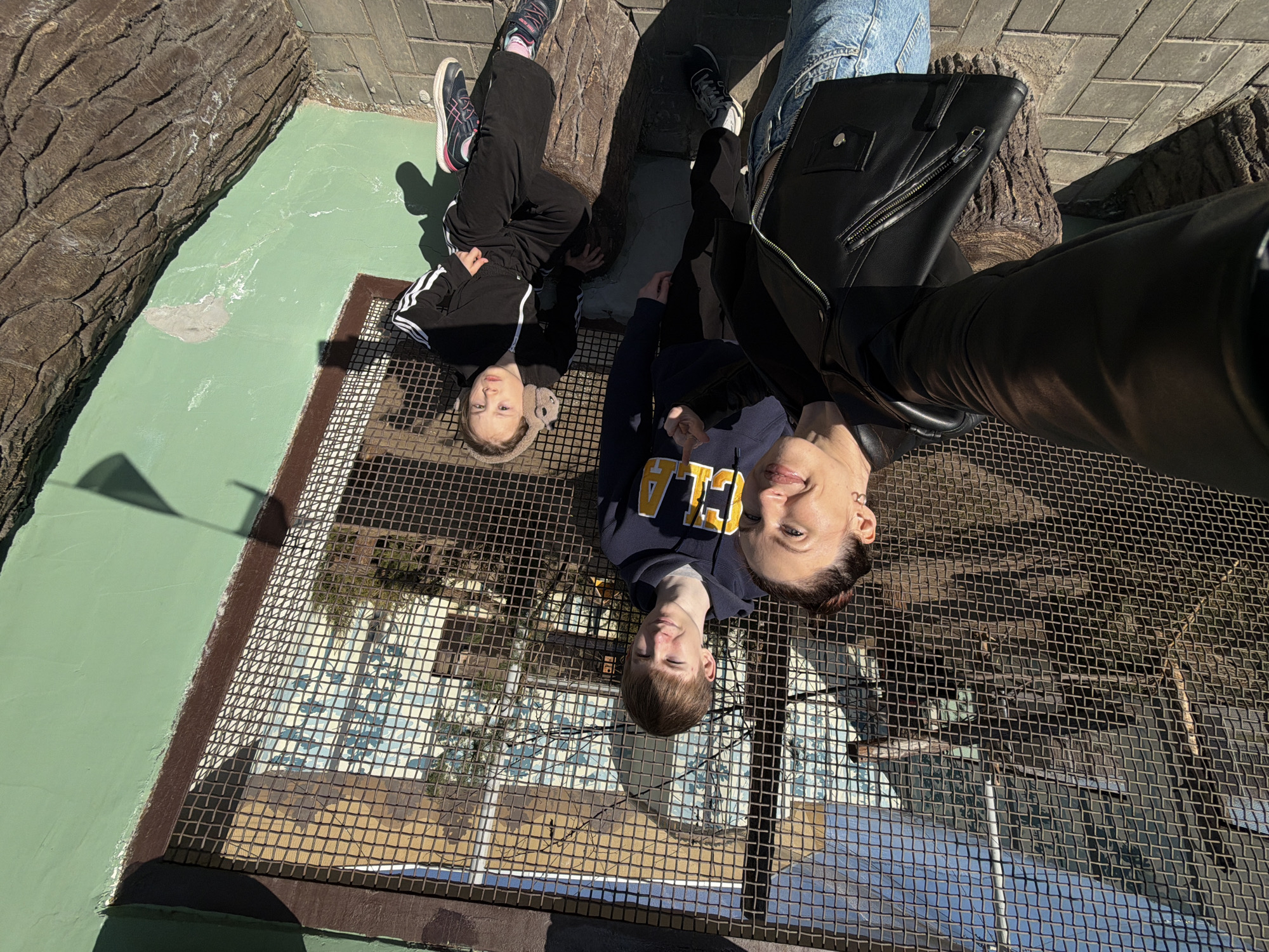 The image shows three individuals photographed from a high angle through a metal grid. The youngest person wears a dark-colored tracksuit with white stripes down the arms and legs, and pink-accented athletic shoes. The middle person wears a dark-colored hoodie with yellow lettering that reads "UCLA". The third person wears blue jeans and a black leather jacket. The people are positioned on the grid above what appears to be a water feature or building visible below. The surroundings include green painted surfaces, faux rock structures and stone-paved areas. The location appears to be the Perm Zoo in Perm, Russia.