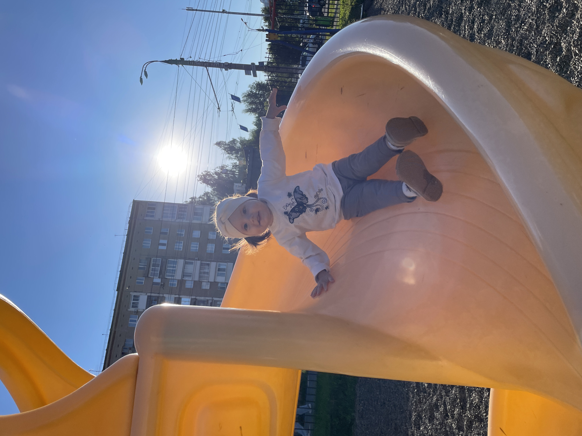 In Izhevsk, Russia, a young child with a light-colored headband and dark hair is positioned on a bright orange playground slide. The child is wearing a white long-sleeved shirt with a butterfly graphic and grey pants. Their feet are extended downwards, suggesting movement on the slide. The background features a blue sky with the sun prominently visible, creating a lens flare. A multi-story apartment building, a street light with wires, and trees are also part of the urban environment. The ground beneath the slide is covered in dark gravel. The image is captured from a low angle, emphasizing the slide and the child's activity.