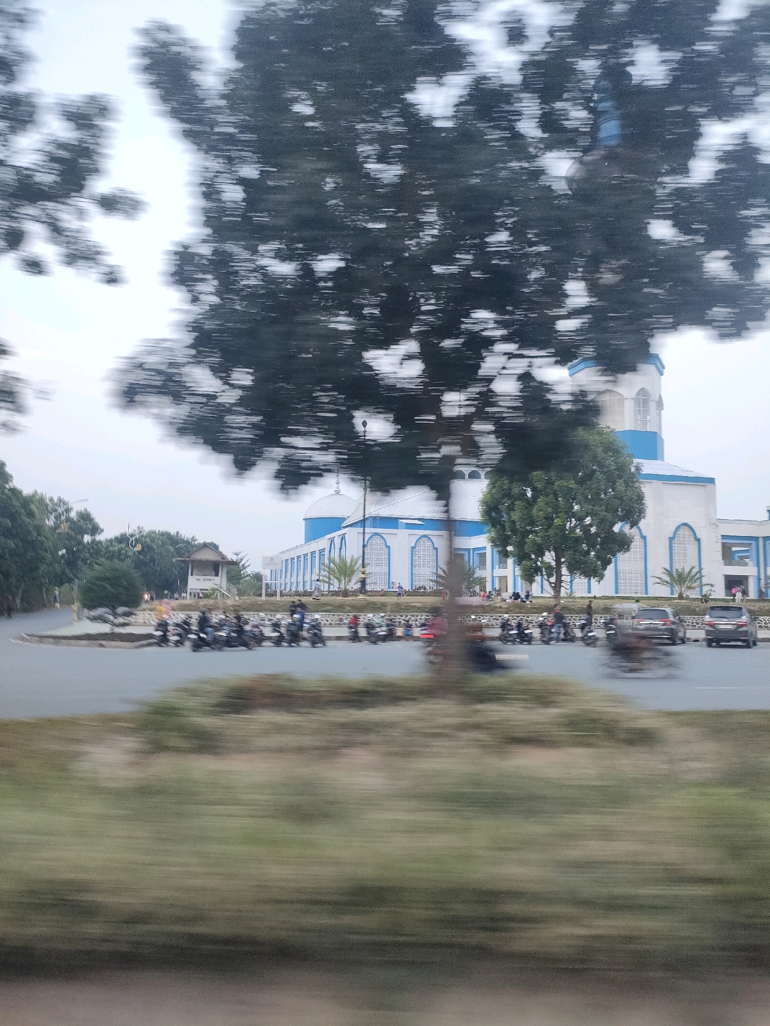 The image depicts a street scene in Batam, Indonesia, featuring a distinctive blue and white mosque in the background. The mosque has a large white dome and blue accents, with arched windows and a tall minaret. In the foreground, there is a road with numerous motorcycles parked and some in motion, indicating active local traffic. Two cars are visible on the road, also blurred by motion. The surrounding environment includes trees, some of which are large and leafy, partially obscuring the mosque. A small, simple building with a triangular roof is situated near the road. The overall impression is of a typical urban street with a prominent religious building and busy local transportation.