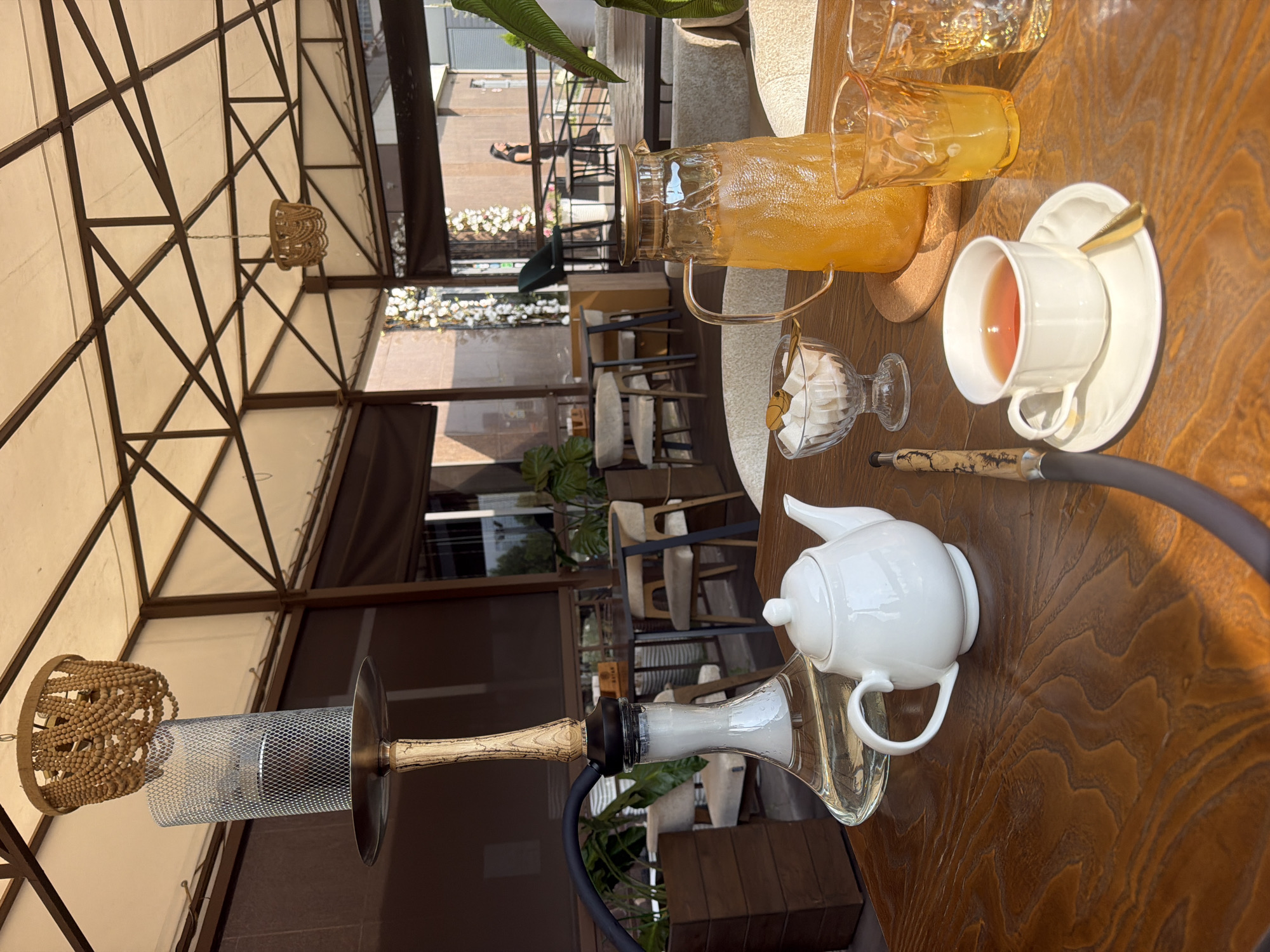 The image depicts an outdoor seating area, likely a cafe or restaurant, on a sunny day. A hookah is prominently displayed on a wooden table in the foreground, with smoke emanating from its base, indicating recent use. Also on the table are a white teapot, a cup of tea, a bowl of sugar cubes, and a pitcher of orange juice with two glasses. In the background, other tables and chairs are visible, along with plants and a woman walking in the distance. The overall ambiance suggests a relaxed setting for socializing or enjoying refreshments. The location is identified as being in Kazan, Russia.