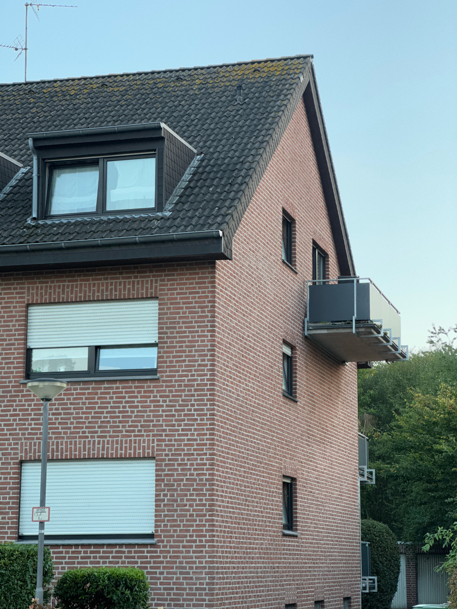 This image shows the exterior of a red brick apartment building in Cleves, Germany. The building has a dark grey tiled roof with a dormer window at the top. There are several windows on the brick facade, some with white roller shutters. A balcony with a metal railing and glass panels extends from the upper floor. Trees and bushes are visible in the background, and a street lamp stands in the foreground. The sky is clear and blue.