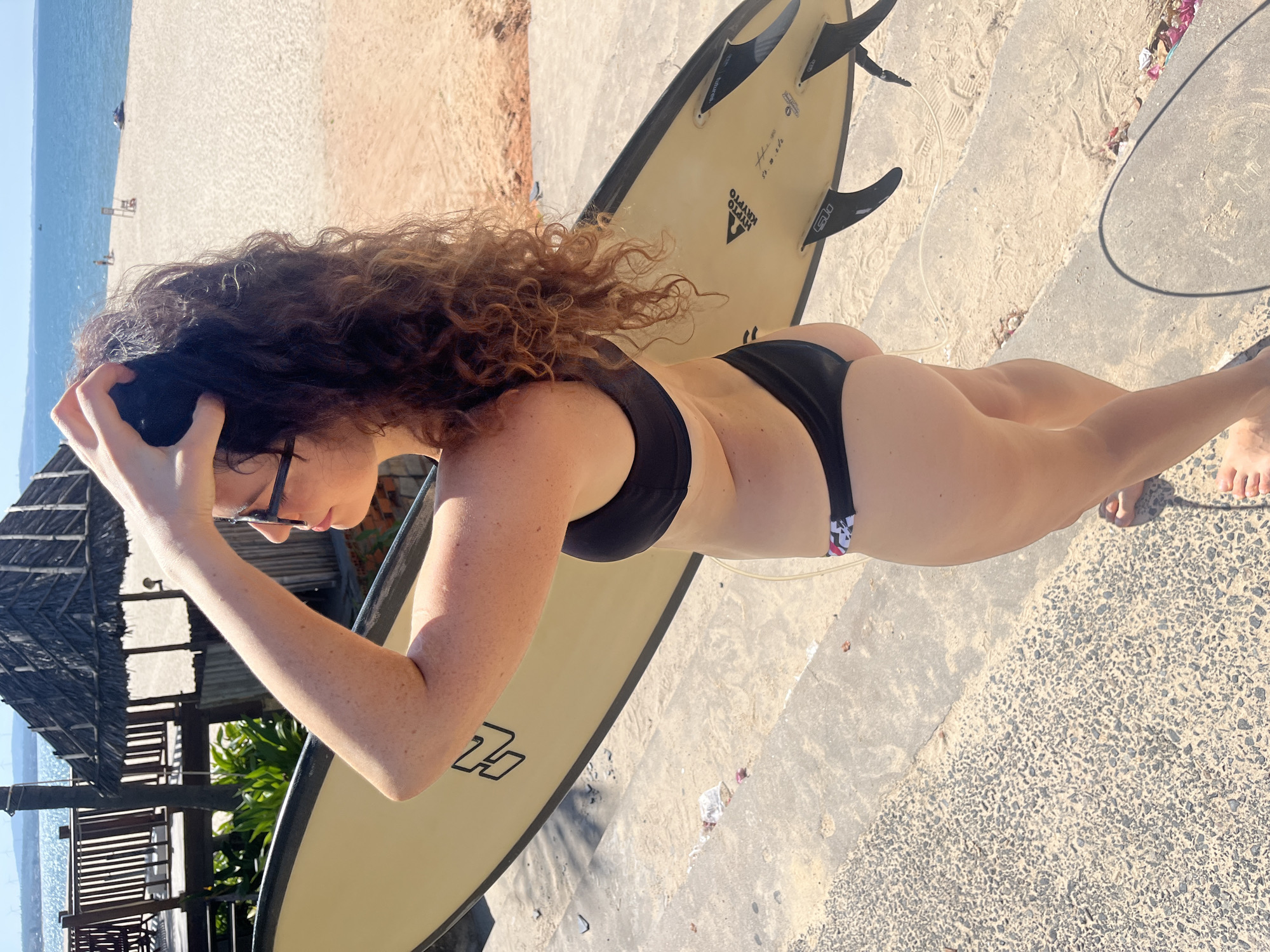 A woman in a black bikini stands on concrete steps near a sandy beach, holding a light-colored surfboard. Her right arm is raised, with her hand in her curly brown hair. She is looking to her left, away from the camera. Behind her, the ocean stretches to the horizon under a clear blue sky. To the left of the woman is a wooden structure with a thatched roof, possibly a beach hut or restaurant, and further back, some trees and what appear to be windmills on a distant hill. The scene suggests a sunny day at a coastal resort in Mũi Né, Vietnam.