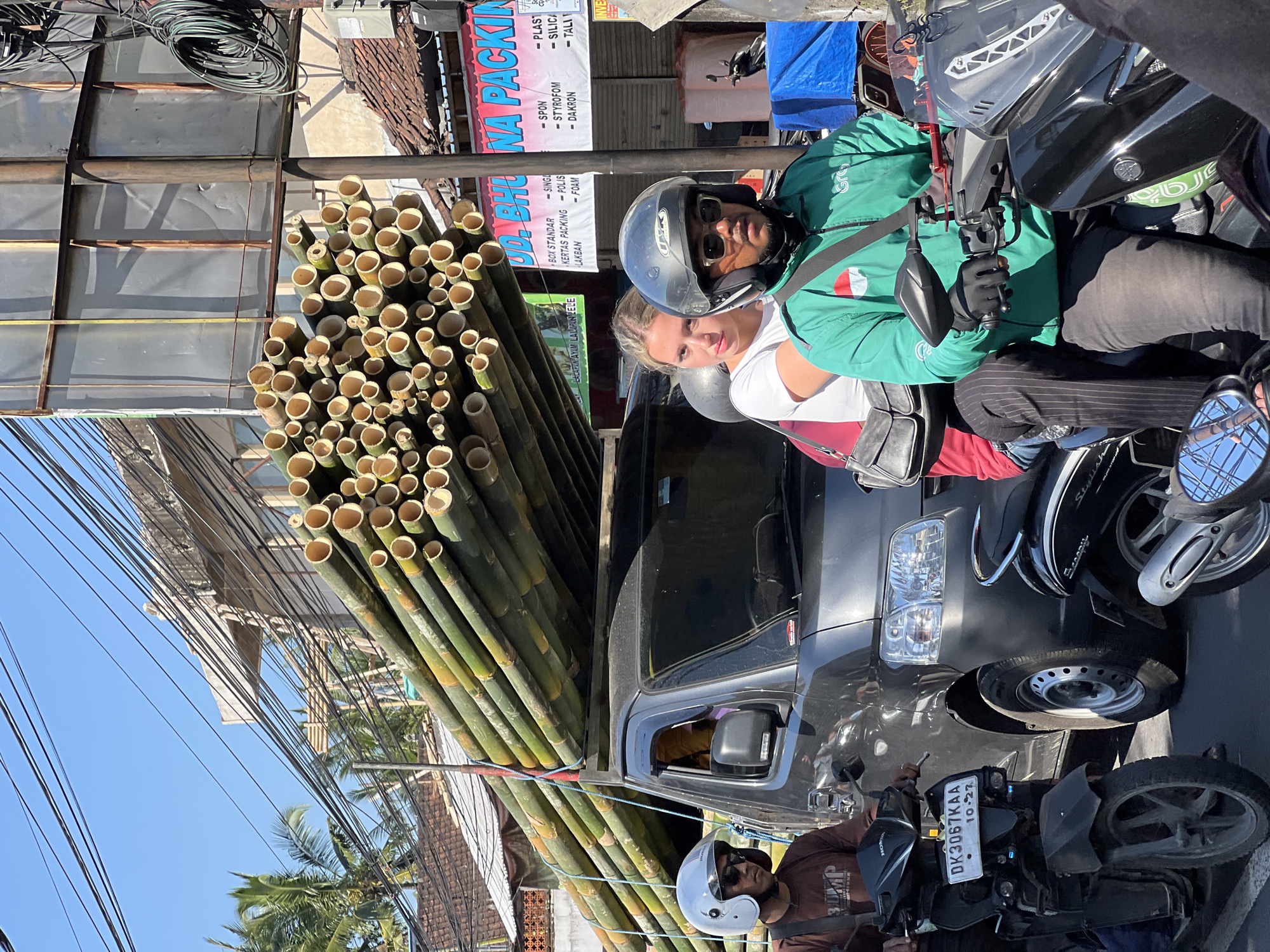 This is a street-level, eye-level, medium shot capturing a scene in the Ubud District, Indonesia, on a sunny day with a clear blue sky. The primary focus is on a dark grey utility vehicle being driven, with a large load of bamboo poles secured on its roof, extending significantly past the vehicle's length. The bamboo is primarily green with some yellowing sections, and the poles are bundled together.
In the foreground, two people are on a motorcycle. The driver, wearing a white helmet, sunglasses, and a brown t-shirt, is on the left. The passenger behind him, a young woman with blonde hair, wears a white t-shirt and a dark purse slung over her shoulder.
To the right of the utility vehicle, another motorcycle is visible. The rider is wearing a black helmet and a bright green jacket with a "Grab" logo and an Indonesian flag emblem. The rider's gloved hands are on the handlebars.
The background reveals typical urban street elements: buildings with tiled roofs, power lines crisscrossing overhead, and a large billboard with faded text that includes "UD. BHU NA PACKING." The overall environment suggests a busy street in a commercial or residential area.