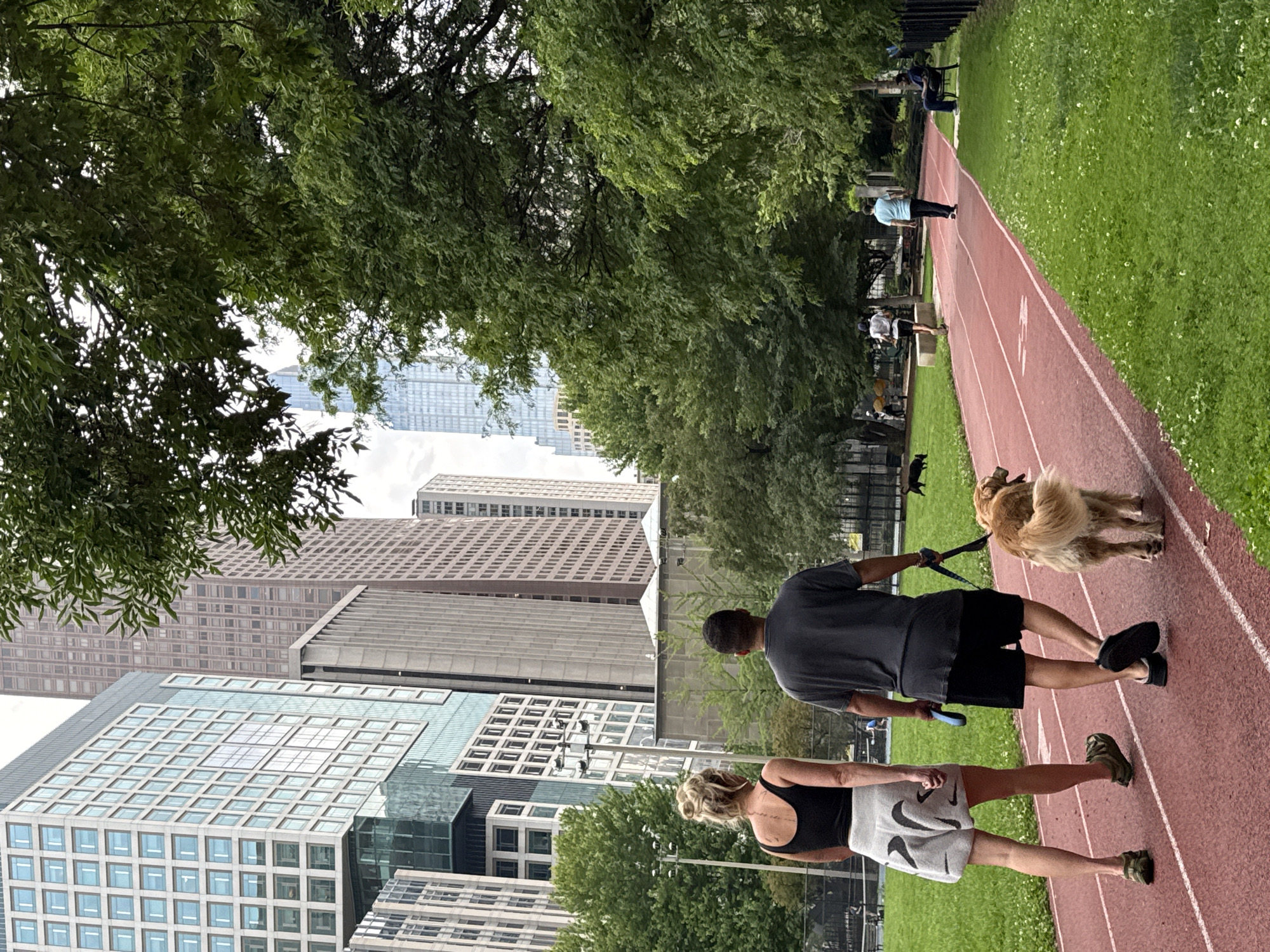 A couple walks their golden retriever on a red running track bordered by green grass and trees. In the background, a row of tall city buildings creates a backdrop. The woman is wearing a black tank top and grey shorts, while the man is in a grey t-shirt and black shorts. The dog is on a leash held by the man. Other people are seen in the distance, some running, one sitting on a bench. The overall scene depicts a park setting within a bustling urban environment, likely in Chicago, United States of America, given the architectural style of the buildings.