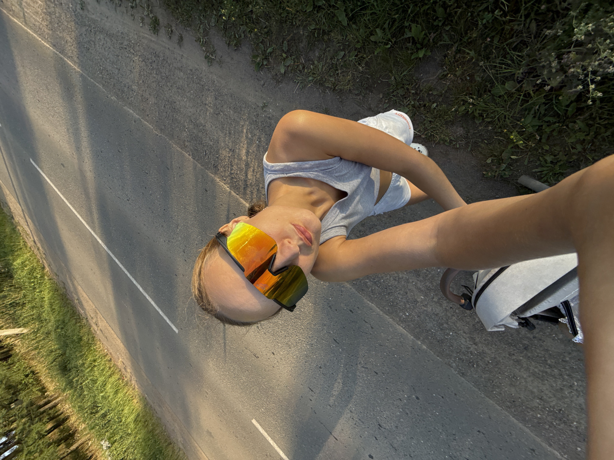 A young woman is captured in a selfie from a high angle, possibly taken while riding a bicycle or motorcycle. She is wearing a grey tank top, white shorts, and mirrored sunglasses with orange and yellow lenses. Her hair is pulled back, and she is looking towards the camera with a neutral expression. The background shows a paved road with a white line, bordered by grass and trees on one side and a dirt shoulder on the other. The lighting suggests it is late afternoon or early evening, with shadows cast across the road. The scene appears to be rural, consistent with a location like Podshivalovo, Russia. The overall mood is casual and outdoorsy. There is visible text on her shorts, which appears to read "UNIVERSITY".