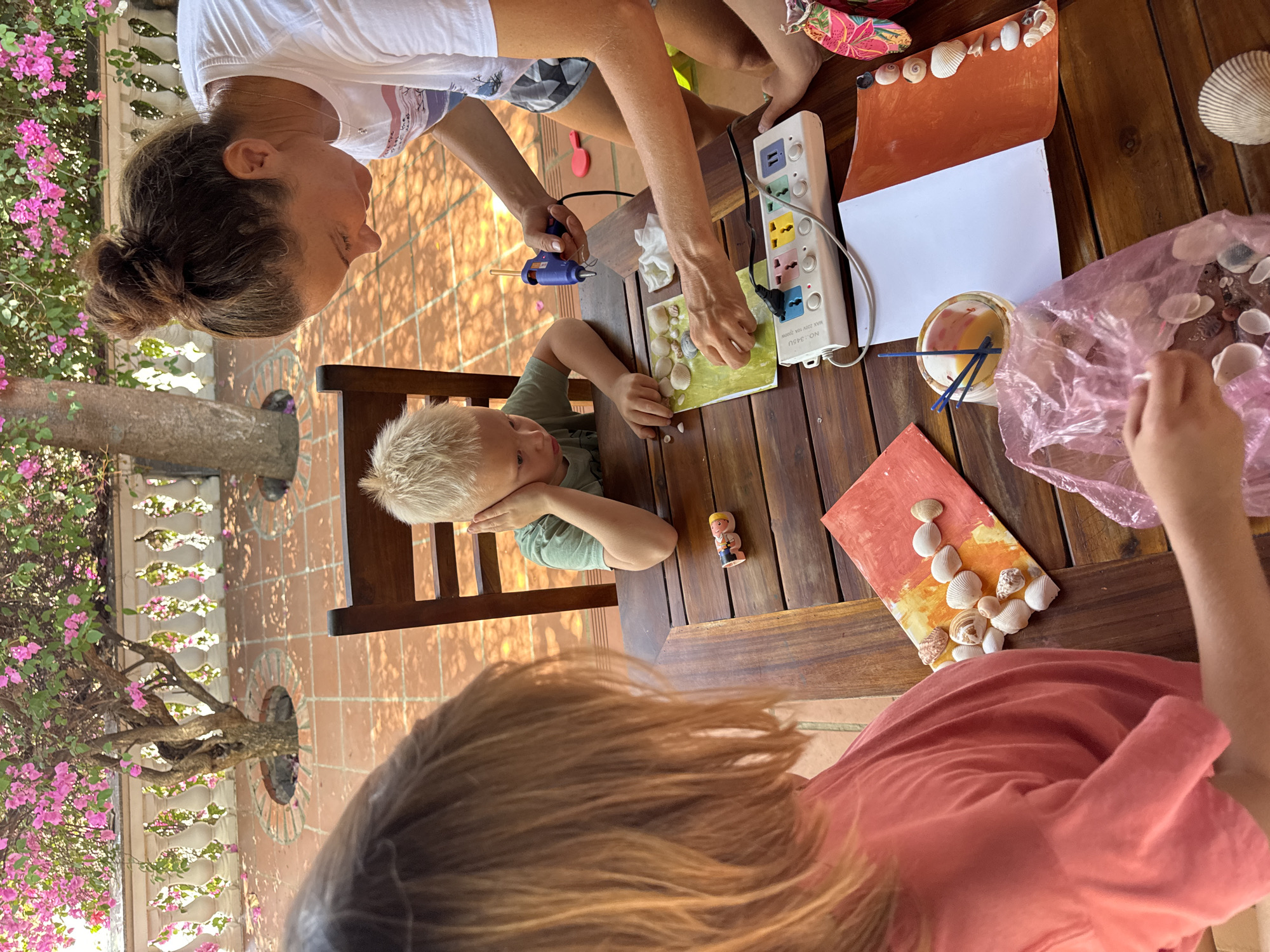 The scene depicts a family crafting on a wooden table, likely outdoors on a patio in Mũi Né, Vietnam, given the tile flooring and nearby bougainvillea. An adult with their hair in a bun is using a glue gun to adhere seashells to a green piece of paper, while a young boy with blond hair watches with his head resting on his hand. Another child, partially visible, is reaching into a pink plastic bag filled with more seashells. Crafting supplies such as paint, brushes, and painted paper are scattered on the table alongside a power strip. A small, toy figurine is on the table as well.