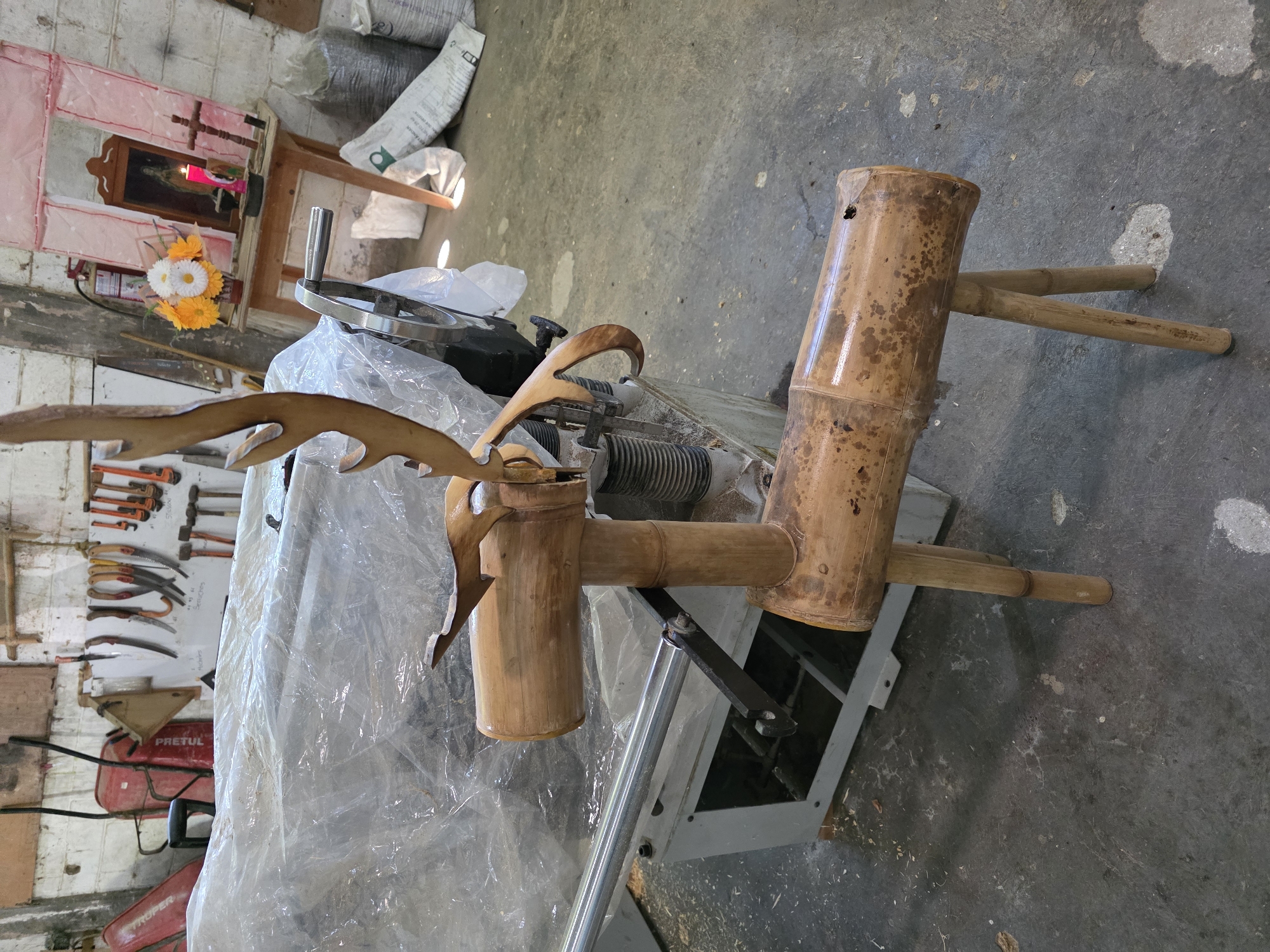 The image depicts a workshop setting in Equimita, Mexico, with a focus on bamboo furniture. The primary subject is a three-legged stool constructed from bamboo, possibly a work-in-progress on a metal-framed machine. A piece of unfinished wood resembling deer antlers is visible near the stool. The background includes a shrine with a religious picture, a lit candle, and artificial flowers, along with bags of unknown materials. A wall displays a collection of tools, including saws, wrenches, and a red wheelbarrow. The environment is cluttered, indicative of a working space.